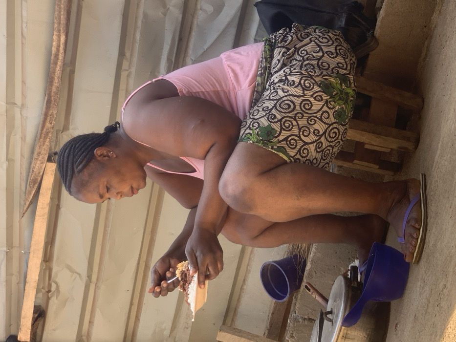 A woman with braided hair and a pink top crouches, eating cake with a fork. She wears a patterned skirt and flip-flops. Nearby are a blue cup and a blue scoop. A cooking pot with a lid and a metal stand are also visible. The background consists of corrugated metal and stacked wooden planks.
