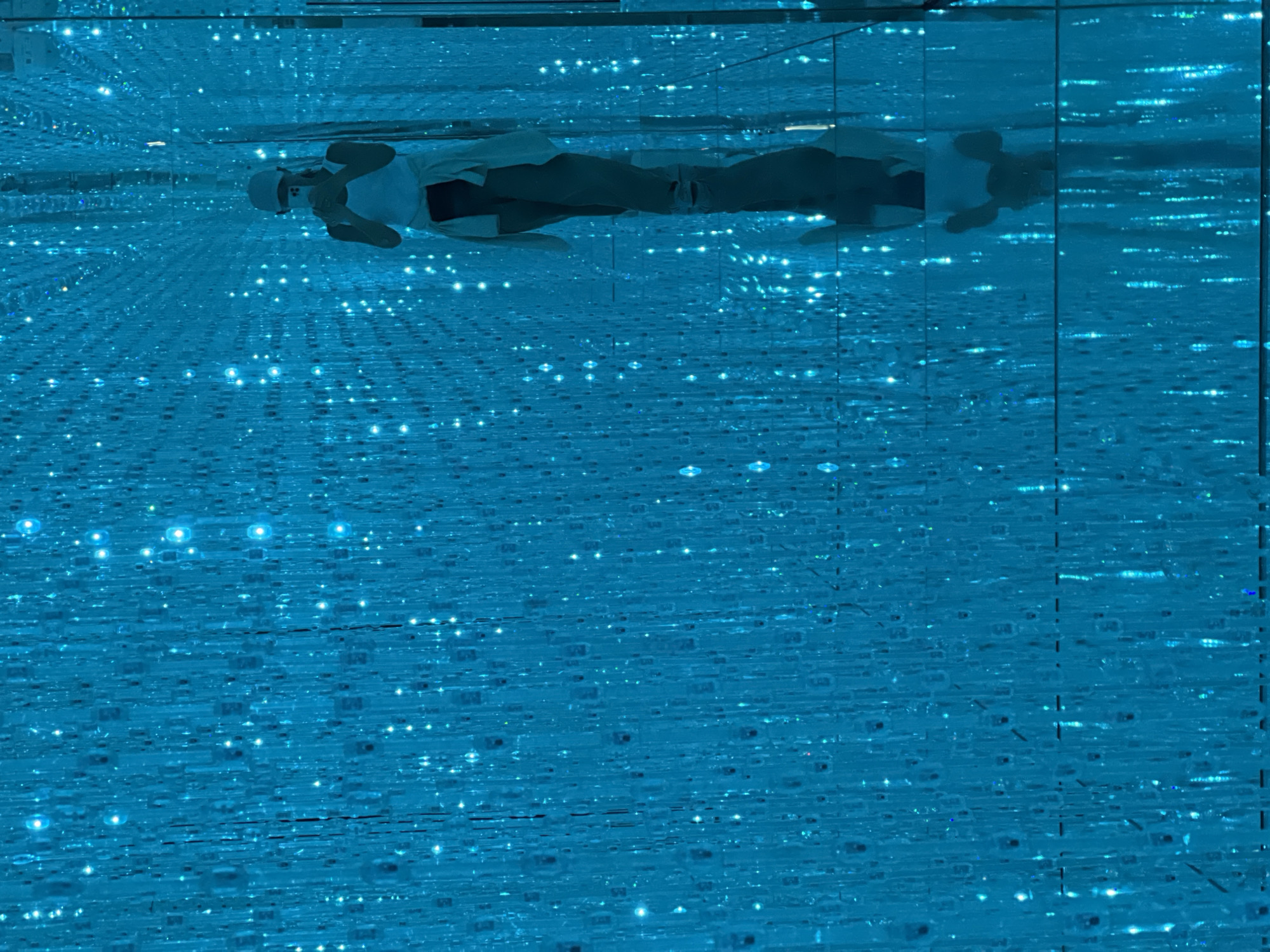 A person is lying down in a room filled with blue lights and mirrors. The person is wearing a white shirt and a white cap, and appears to be lying on their back. Their reflection is visible in the mirrors, creating an illusion of depth and repetition. The room is dark except for the numerous small blue lights scattered throughout, which are also reflected in the mirrors, giving the impression of a starry sky or a digital landscape. The overall atmosphere is serene and immersive. The image appears to be taken indoors, in a controlled environment. There is no indication of the time of day, weather, or specific location beyond the general context provided. No text is visible in the image.