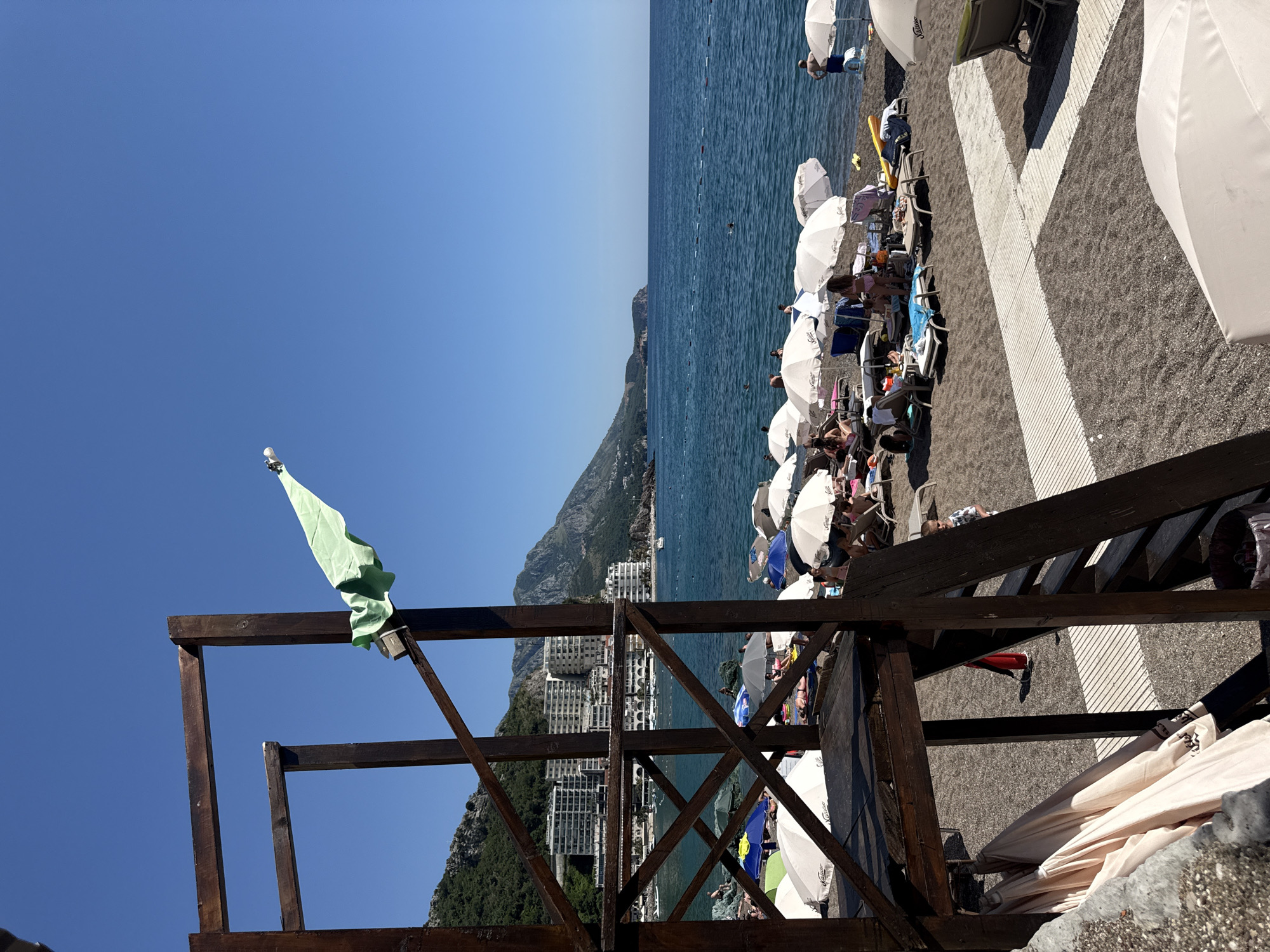 This image depicts a sunny beach scene in Rafailovići, Montenegro. In the foreground, a dark brown wooden multi-level structure, resembling an observation or lifeguard tower, occupies the left side. A light green piece of fabric, possibly a towel or flag, hangs draped over an outward-extending wooden beam on the upper right side of this structure, positioned against the clear blue sky. Below the structure's main platform, several folded beige beach umbrellas are stored. A partial view of a gray concrete wall is visible in the bottom left.
The beach itself is composed of dark gray pebbles or coarse sand. Numerous white beach umbrellas, along with a few colored ones (blue, green, gray), are set up across the midground, accompanied by lounge chairs. Many individuals are present, engaging in typical beach activities; some are reclining under umbrellas, others are standing near the water's edge. A small child in a striped shirt is visible near the stairs of the wooden structure. White slatted wooden pathways traverse the pebble beach. A red trash can is partially visible near the wooden structure, and a yellow inflatable object lies on the sand near the water.
The calm, deep blue-green sea extends across the middle and right sections of the image. A line of orange buoys marks a designated swimming area, where several people are seen swimming or wading. In the background, lush green mountains rise steeply, dotted with numerous white multi-story buildings, likely hotels or apartments, featuring balconies. The coastline recedes to the right, showing a distant headland. The sky is uniformly clear and bright blue.