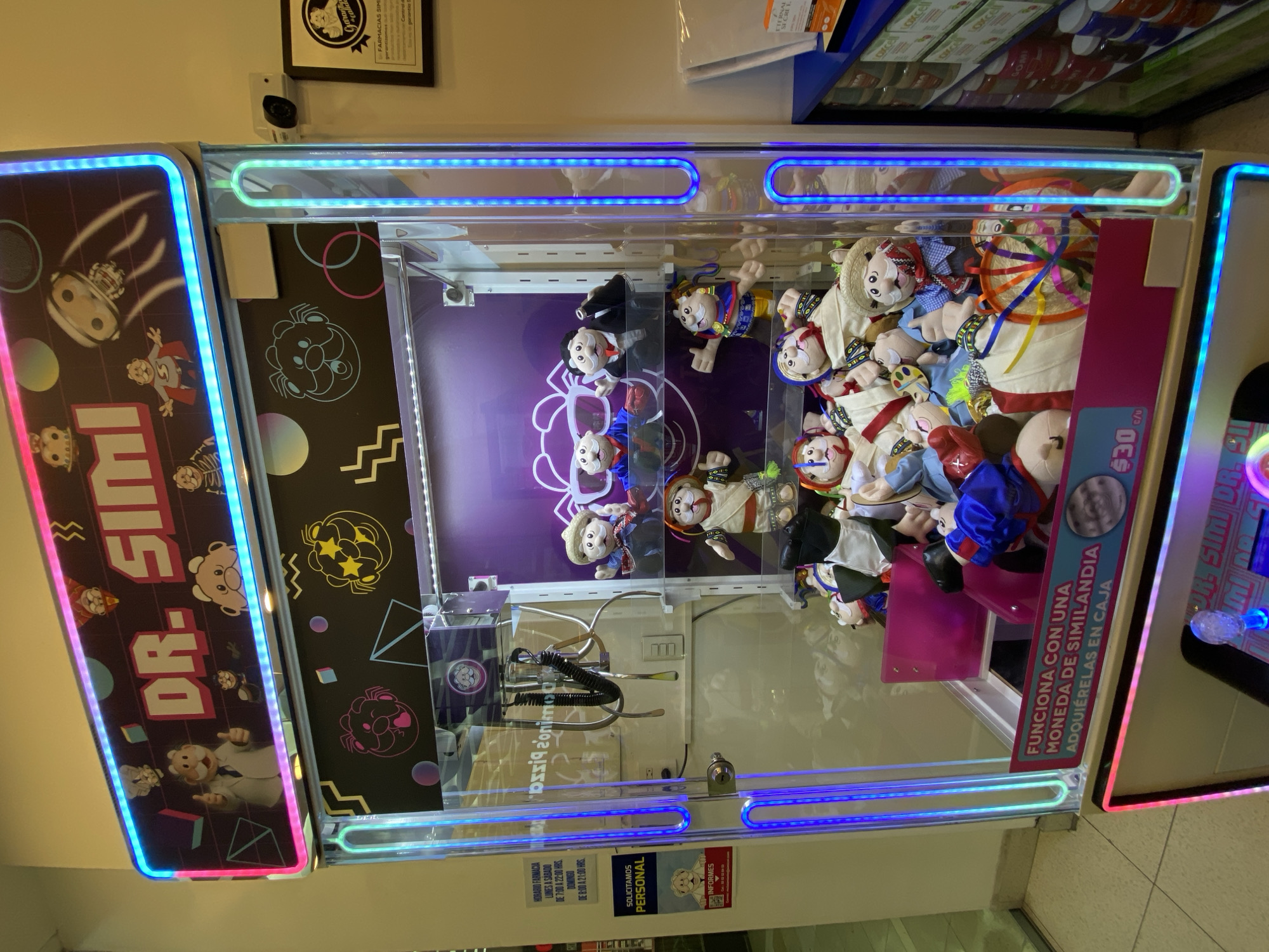 The image shows a Dr. Simi claw machine, a type of arcade game, likely located in San Bernardino Tlaxcalancingo, Mexico. The machine is filled with plush Dr. Simi dolls, the mascot of a Mexican pharmaceutical chain. The machine's signage indicates that it costs 30 pesos to play and that it accepts "Similandia" coins, which can be purchased at the counter. The claw mechanism is visible inside the machine. The exterior of the machine features "DR. SIMI" branding and colorful light strips. Additional shelves stocked with products are visible above the machine. The area appears to be a brightly lit retail space. Signage indicating pharmacy hours and personnel requests are visible on the machine.