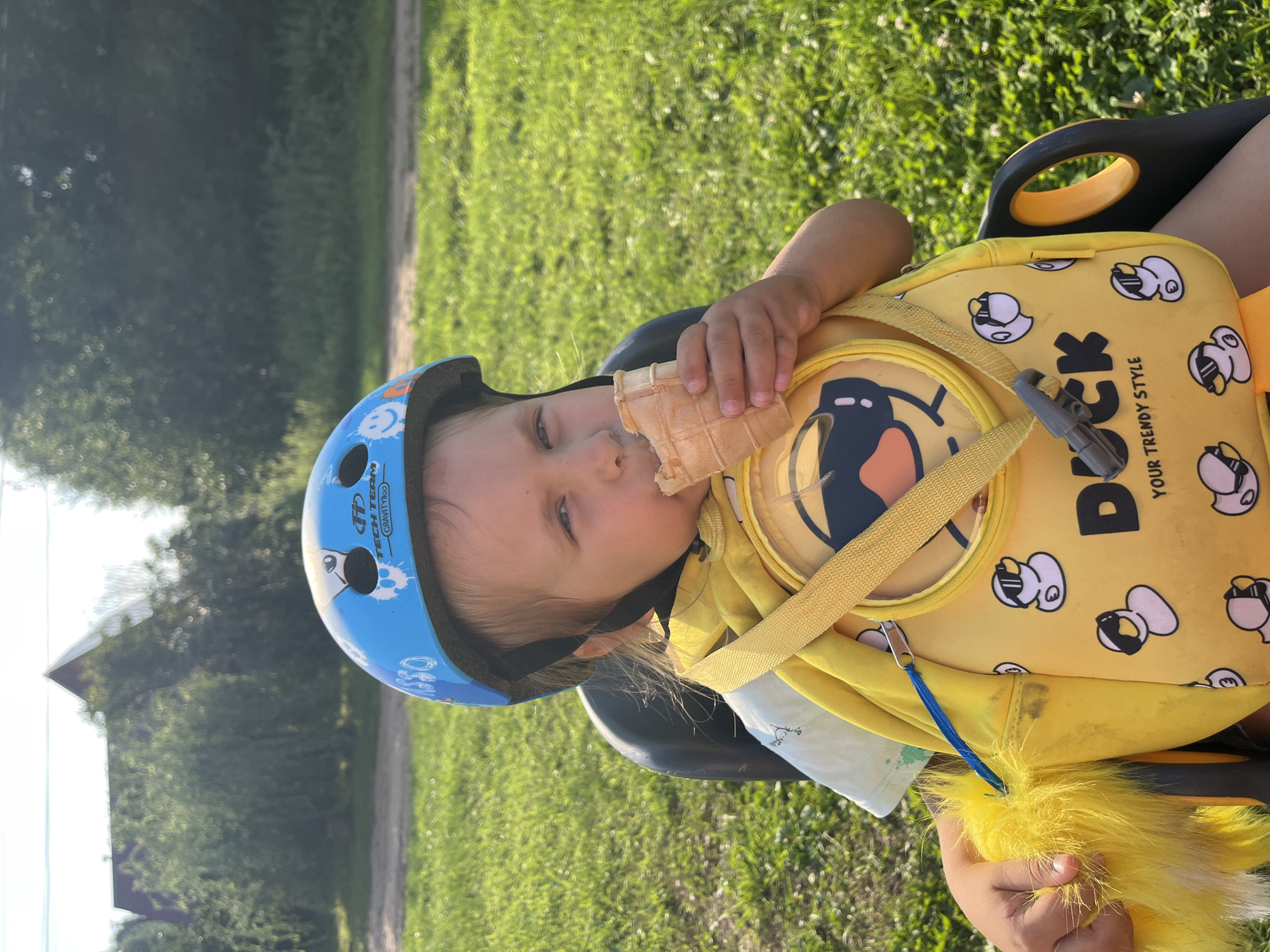 A young child wearing a blue helmet is seated in a black plastic child seat attached to a bicycle, in an outdoor grassy area. The child is holding a partially eaten ice cream cone in a waffle cone and bringing it to their mouth. The child is also wearing a yellow backpack with a duck design. The background shows a dirt path, green grass, trees, and a building with a red roof, characteristic of a rural or suburban setting in Chaikovsky, Russia. The lighting suggests it is daytime with sunlight.