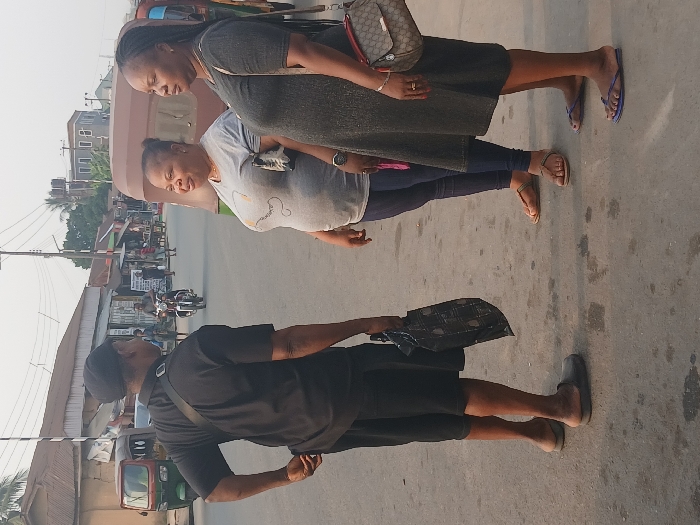 The image displays three individuals on a paved road in Aba, Nigeria, engaged in a conversation under daylight conditions. On the left, a male is seen from behind, wearing a black cap, black short-sleeved shirt, black shorts, and dark slip-on shoes, with a dark strap across his chest and holding a dark, patterned bag in his right hand. He faces two females standing to his right. The first female, positioned centrally, wears a light grey t-shirt with a dark graphic, dark blue leggings, and light brown flat sandals; she exhibits a slight smile. The second female, on the far right, wears a dark grey knee-length dress and blue flip-flops, carrying a brown and beige patterned crossbody bag on her right shoulder; her expression is neutral.
The background reveals a street lined with various commercial and residential buildings, including single-story structures with corrugated metal roofs and taller buildings in the distance. Utility poles with overhead wires are present. Vehicles include a green and red motorized rickshaw (keke Napep) on the far left, another brown motorized rickshaw partially visible behind the women, and a person riding a motorcycle further down the street. The road surface is asphalt with visible stains or patches.