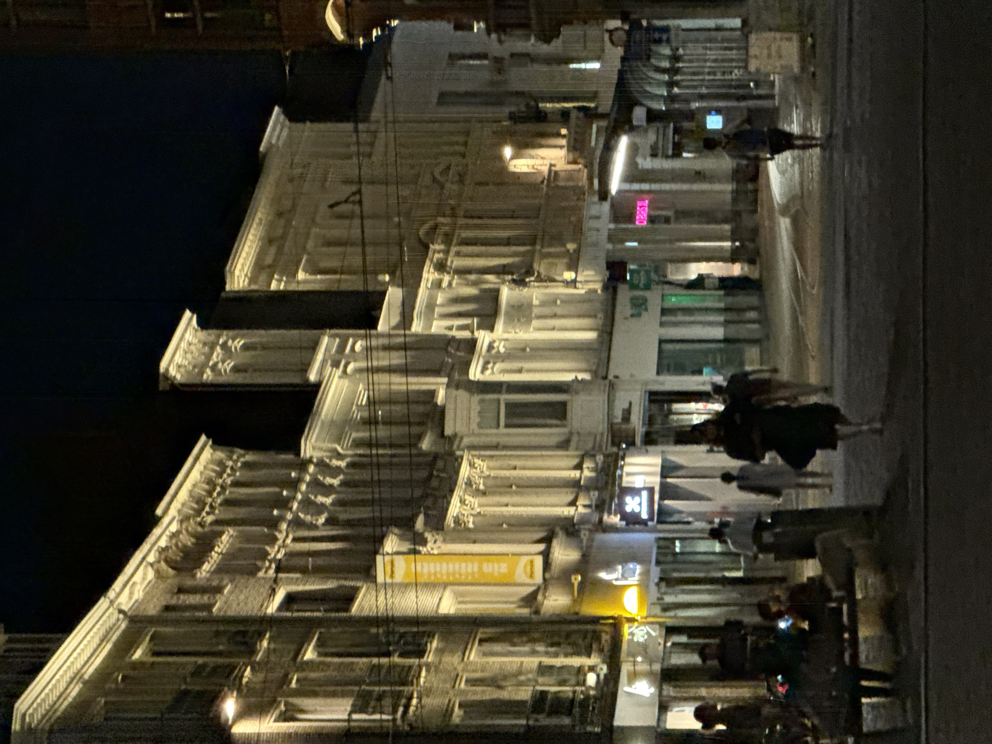The image depicts a street scene at night in Ghent, Belgium, possibly near the Veldstraat shopping area, known for its historical architecture and shops. The focus is on the illuminated facades of buildings lining the street. A variety of shops are visible, with some displaying bright signage, including a yellow banner with text. Pedestrians are present on the sidewalk, some walking and others seated on a bench. The lighting suggests a commercial area, with streetlights and shop lights casting a warm glow on the scene. The architecture appears to be a mix of older and more modern styles. The overall atmosphere is calm and urban.