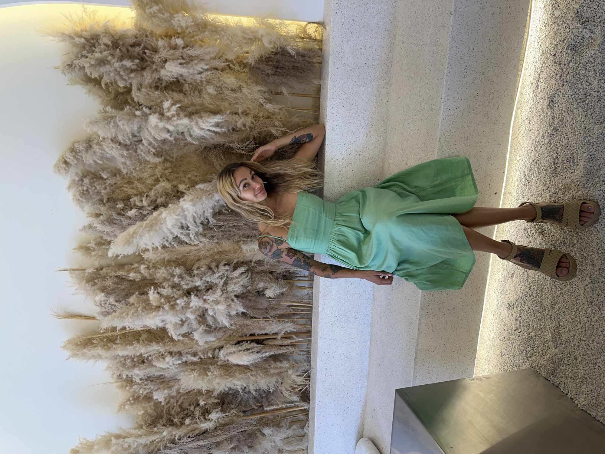 A woman lies in Phu Thuy Ward, Vietnam, wearing a green strapless dress and brown sandals. She has blonde hair and tattoos on her arms. She is positioned between a wall of dried pampas grass and a light-colored ledge. A section of gravelly ground is visible near her feet, and a corner of a gray structure is in the foreground.