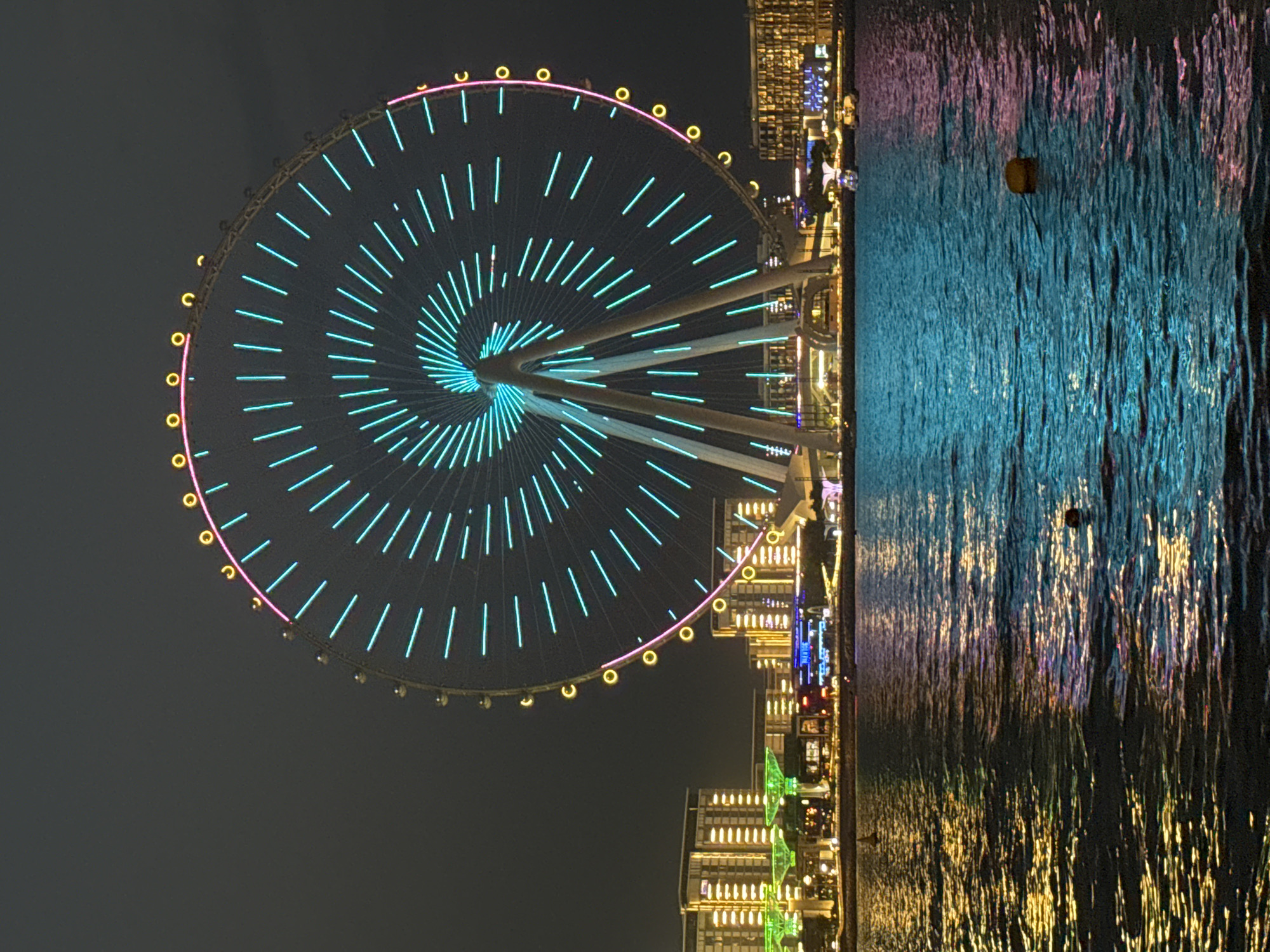 This is a nighttime, eye-level, slightly tilted medium shot of the Ain Dubai observation wheel, the largest in the world, situated on Bluewaters Island in Dubai, UAE. The massive Ferris wheel is adorned with thousands of LED lights that glow in vibrant shades of turquoise and pink, creating a dazzling spectacle against the dark sky. The wheel's structure, including its spokes and the outer rim with its passenger cabins, is clearly visible. The water in the foreground reflects the bright lights of the Ferris wheel and the surrounding cityscape, creating a shimmering, colorful effect on the surface.
On the right side of the frame, the illuminated buildings of Bluewaters Island rise up, showcasing a modern architectural style with a multitude of windows and lights. The reflections in the water suggest a calm, still body of water, likely the Persian Gulf. A small, yellow buoy is visible in the water. The overall scene conveys a sense of urban vibrancy and advanced engineering, typical of Dubai's skyline. The time of day is clearly night, and the weather appears clear, allowing for excellent visibility of the illuminated attractions. There is no visible text in the image.