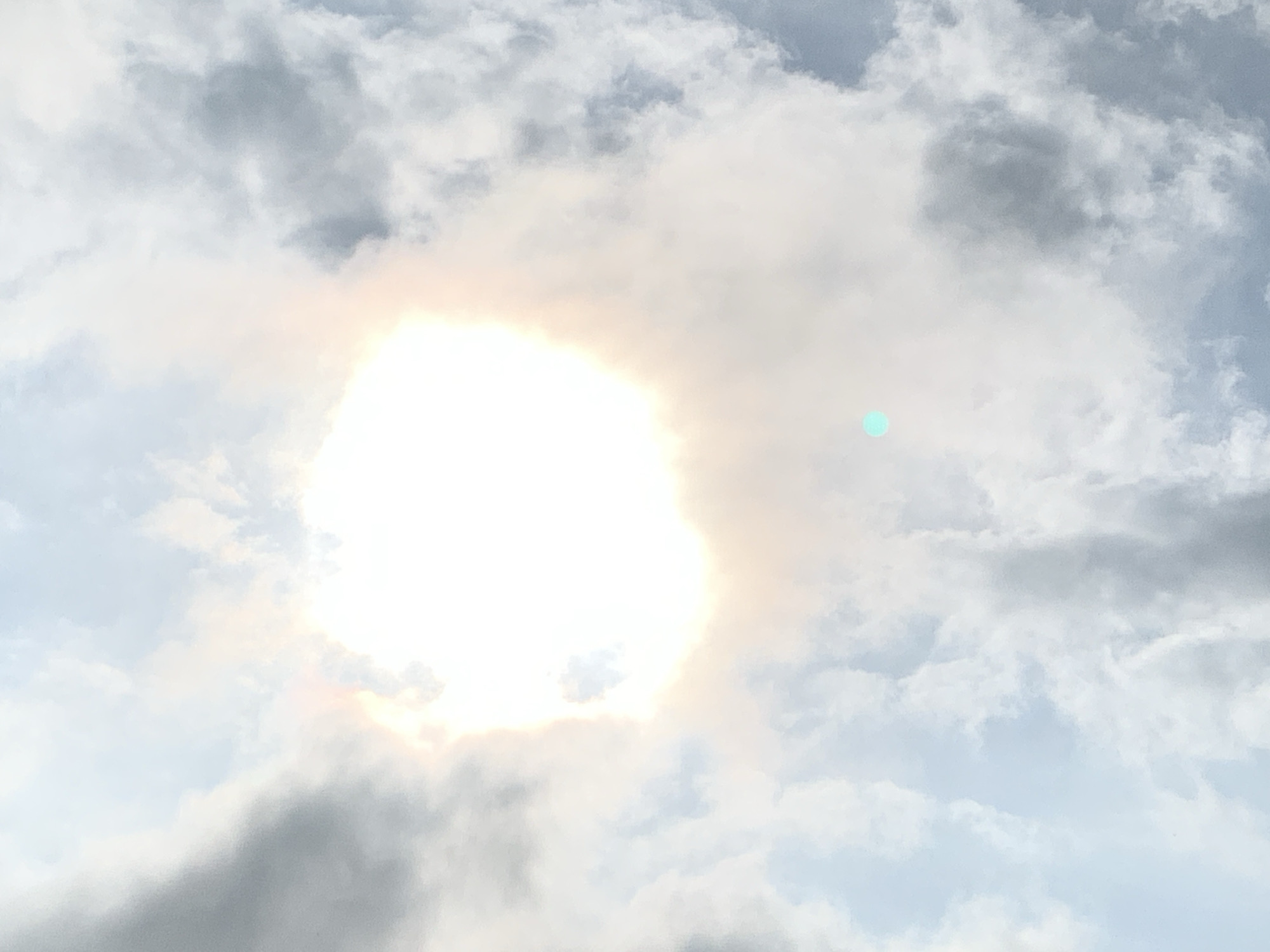 The image shows a bright, partially obscured sun with light rays extending outwards, breaking through a dense cloud cover. The clouds are a mixture of white and grey, with some areas appearing lighter and more diffused, suggesting either sunlight filtering through or a hazy atmosphere. The sky is a pale blue in the background. There is a small, teal-colored circular anomaly visible to the right of the sun, possibly a lens flare or artifact. The scene appears to be an outdoor environment, likely a sky view without any discernible human presence or city structures. The location in Kuru Karama, Nigeria, is not visually identifiable from the provided image alone.