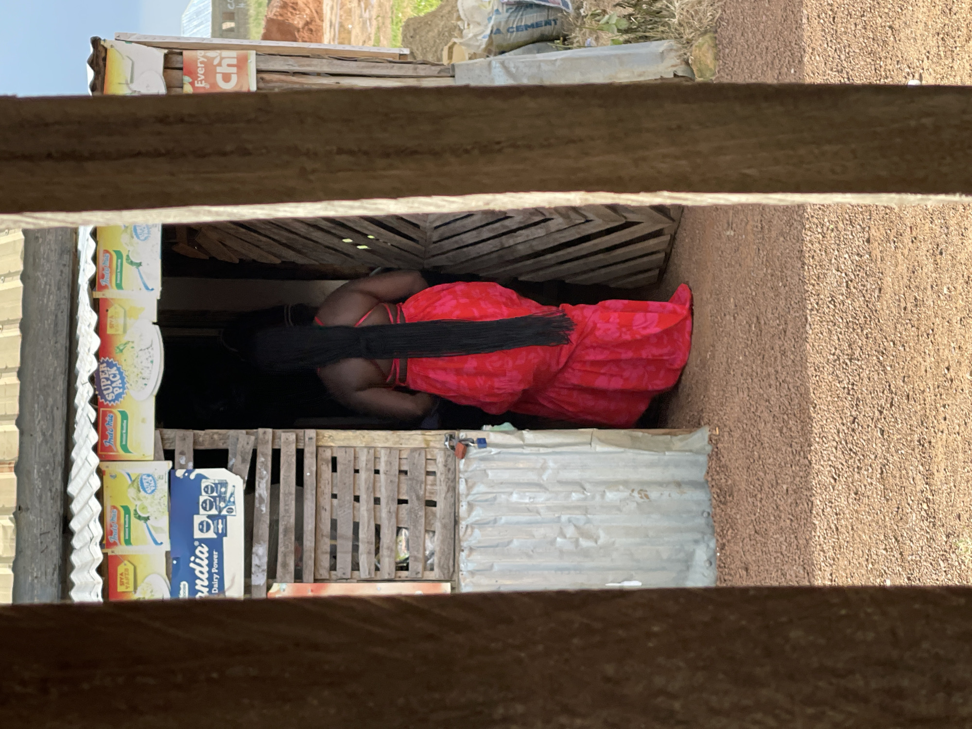 In Durumi, Nigeria, a person stands with their back to the camera, wearing a vibrant red dress with a floral pattern. Their long, dark hair cascades down their back. They are positioned at the entrance of a small, open-air shop, the structure of which appears to be made of corrugated metal and wood. The shop's interior is dimly lit and partially obscured. Visible on the front of the shop are advertisements for "Indomie Super Pack" noodles and "Ondia Dairy Power" milk. The ground in front of the shop is covered in gravel. The environment outside the shop suggests a rural or developing urban setting, with construction materials, including bags of cement, visible in the background. The scene is framed by two vertical wooden posts in the foreground, suggesting the viewer is looking through an opening.