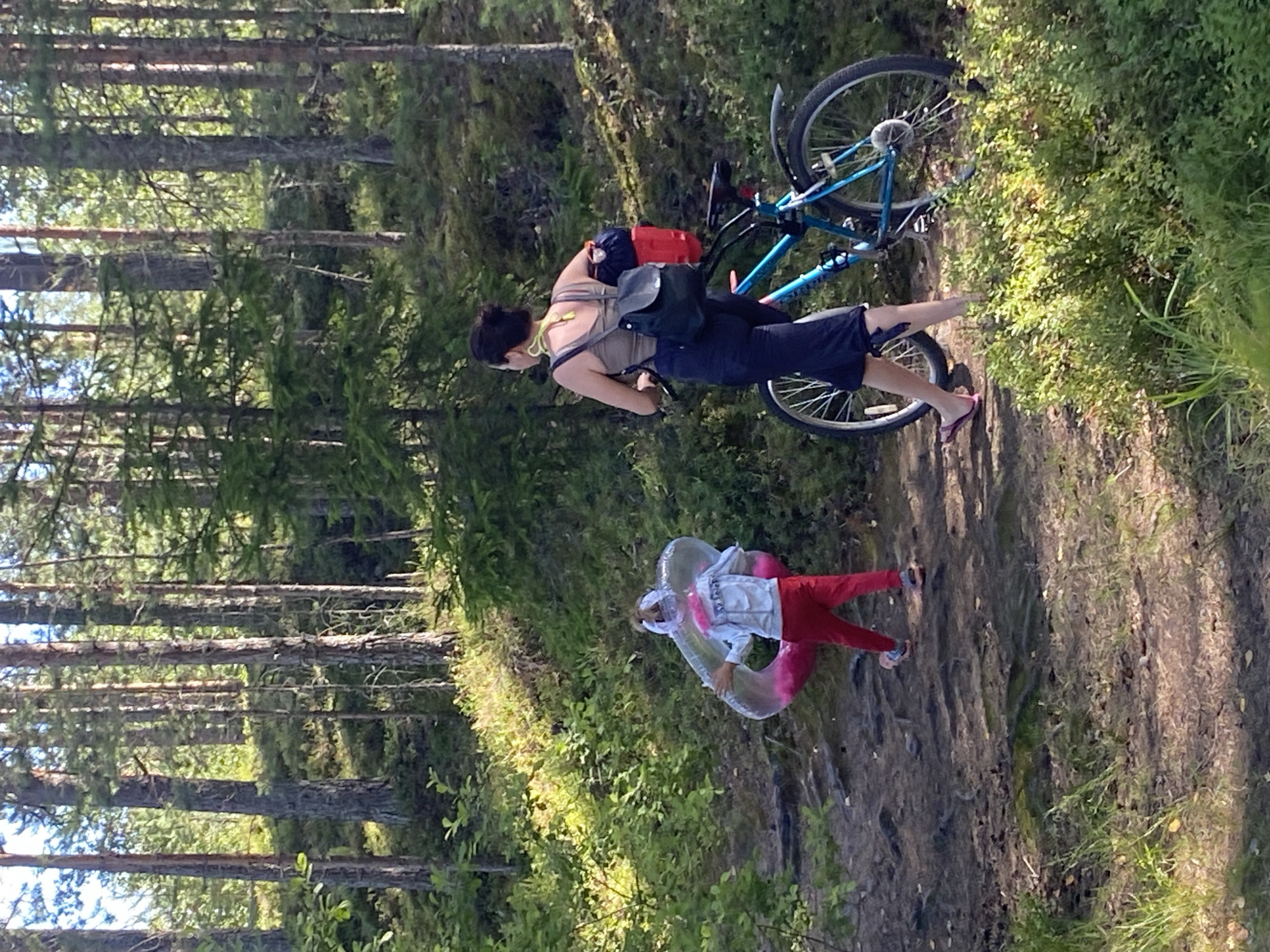 In a sun-dappled forest in Leipyasuo, Russia, a woman on a blue bicycle, laden with a red and blue pack, pauses on a dirt path. She wears dark blue pants and a beige tank top. Ahead of her, a child in red pants and a light-colored jacket walks away, holding a large, inflatable pink and clear ring. The path is uneven, covered with dirt, roots, and scattered leaves, winding through a dense pine forest with undergrowth of green bushes and moss. The light filters through the tall trees, casting shadows on the forest floor, indicating it is likely daytime.