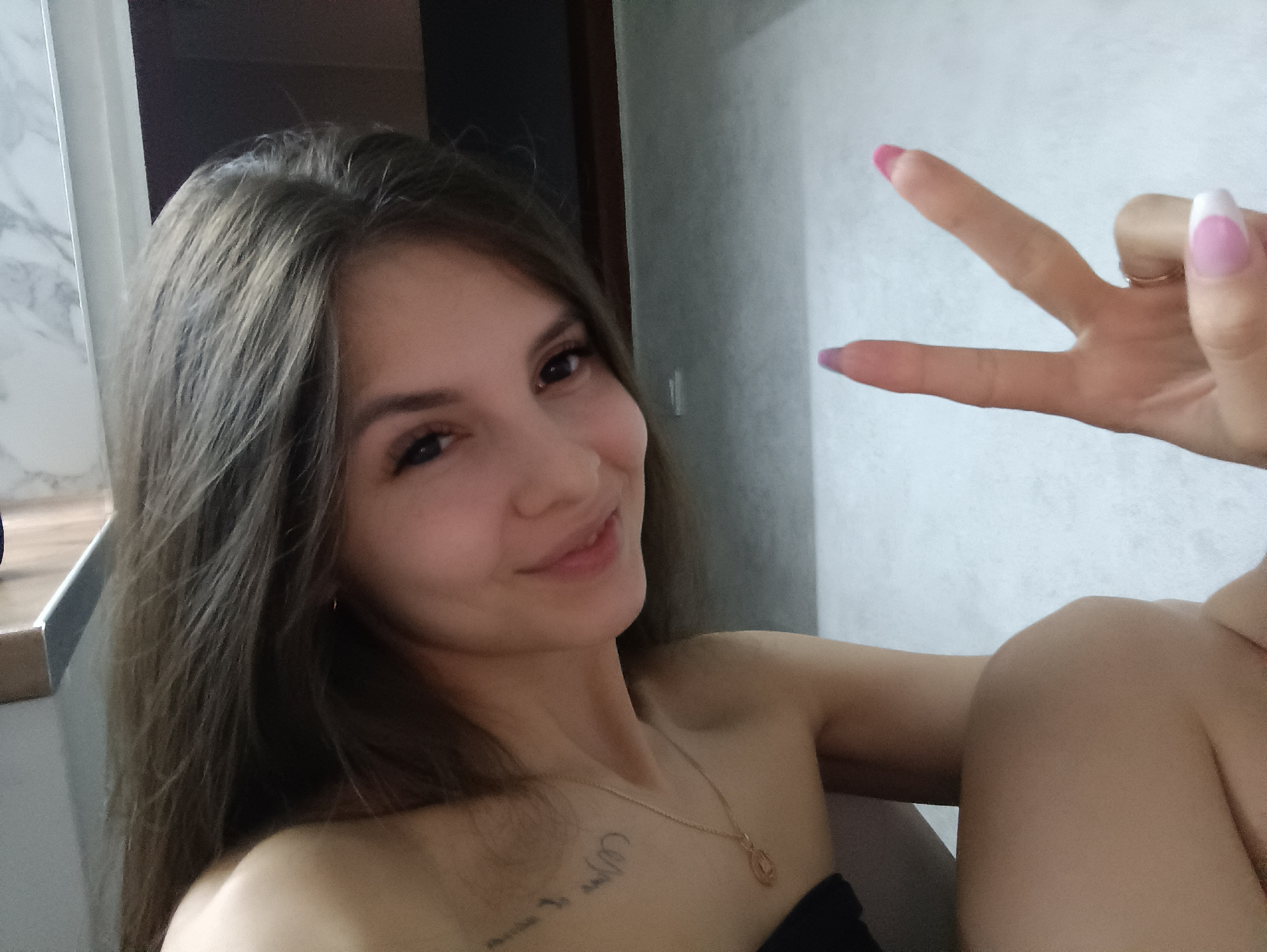 A young woman poses for a selfie in an indoor setting. She is smiling and making a peace sign with her right hand. She has long, dark brown hair and appears to be wearing a tube top. A gold necklace with a pendant is visible around her neck. There is a tattoo visible on her chest. The background includes a light-colored wall, a doorway, and a section of what seems to be a marble countertop. The location is specified to be Chaikovsky, Russia.
