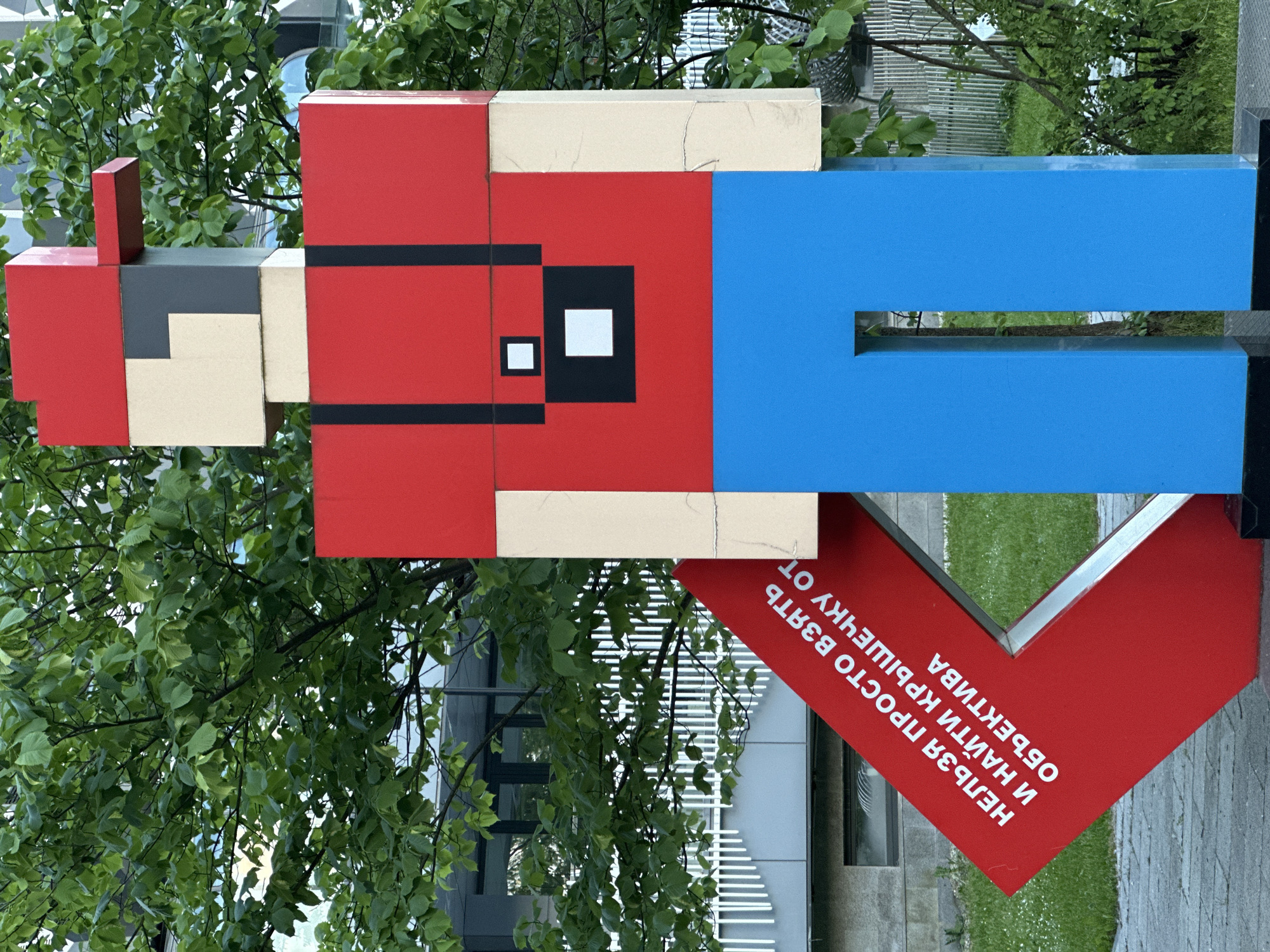 An outdoor, eye-level shot captures a large, blocky, three-dimensional sculpture resembling an 8-bit video game character. The figure, oriented vertically, is composed of geometric shapes in bright red, blue, beige, and black. Its torso is red with black horizontal stripes and a stylized black eye featuring a white square pupil. The lower body is blue, and the head is primarily beige with a gray section and a small red block atop it, suggesting a hat.
Below the figure and angled towards the viewer is a large, glossy red sign shaped like a heart. It bears white Russian text that reads: "НЕЛЬЗЯ ПРОСТО ВЗЯТЬ И НАЙТИ КРЫШЕЧКУ ОТ ОБЪЕКТИВА" which translates to "You can't just take and find a lens cap from a lens." This phrase likely serves as a playful or philosophical commentary.
The sculpture is set against a backdrop of lush green trees with dense foliage, suggesting a park or urban green space. In the background, reflections of modern buildings with linear patterns are visible on glass surfaces, indicating an urban environment in Moscow, Russia. The scene is well-lit, suggesting daytime under clear weather conditions. The overall impression is one of a public art installation with a humorous or thought-provoking message.