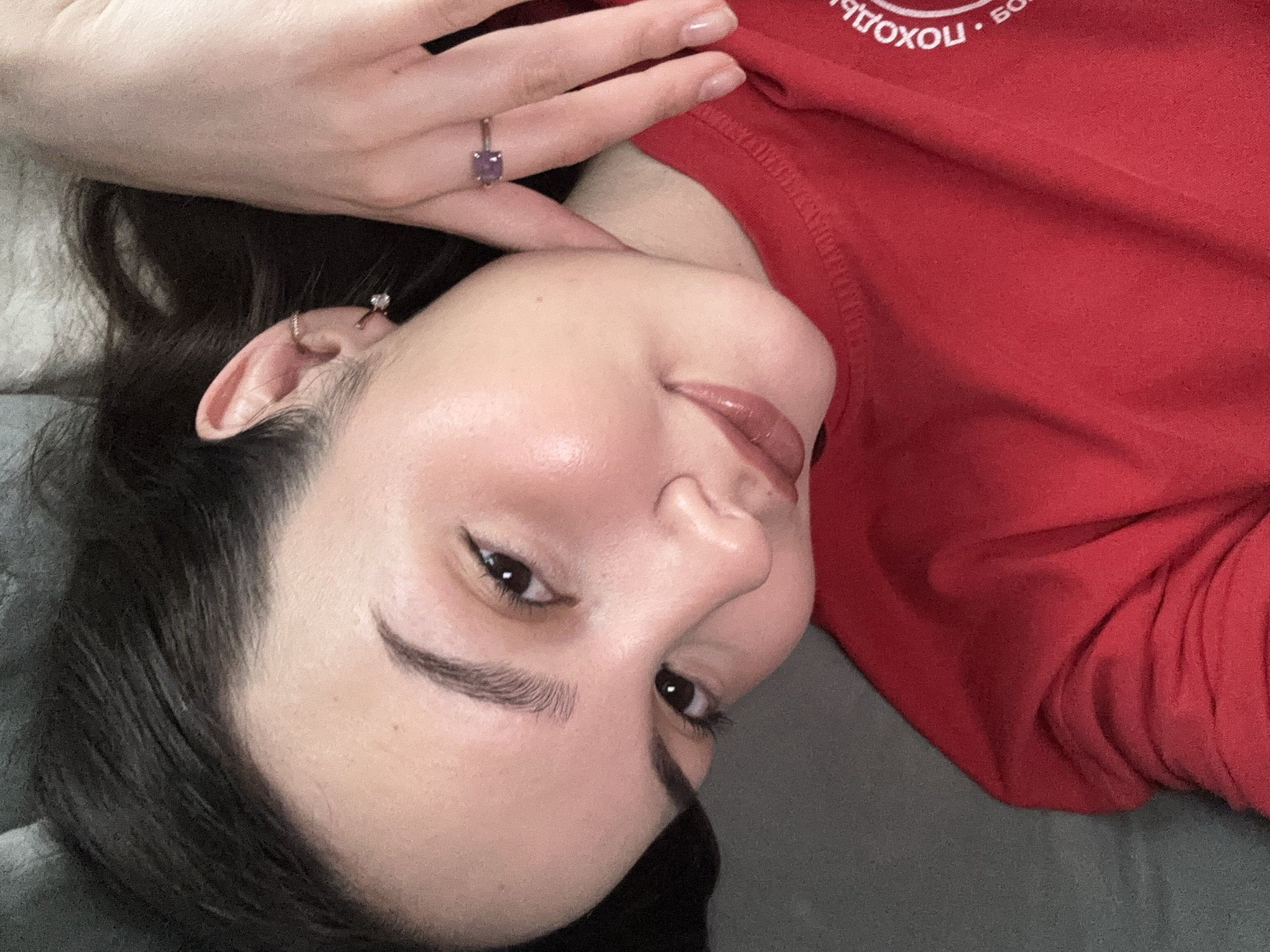 A young woman with dark hair and fair skin is lying down, face angled towards the camera. She is wearing a red sweatshirt with Cyrillic text partially visible. A ring with a purple stone is visible on her left hand, which is resting near her face. She has dark eyebrows and brown eyes, and her lips are glossy. The background appears to be a gray surface. It is not possible to verify the location as Chaikovsky, Russia, without additional information.
