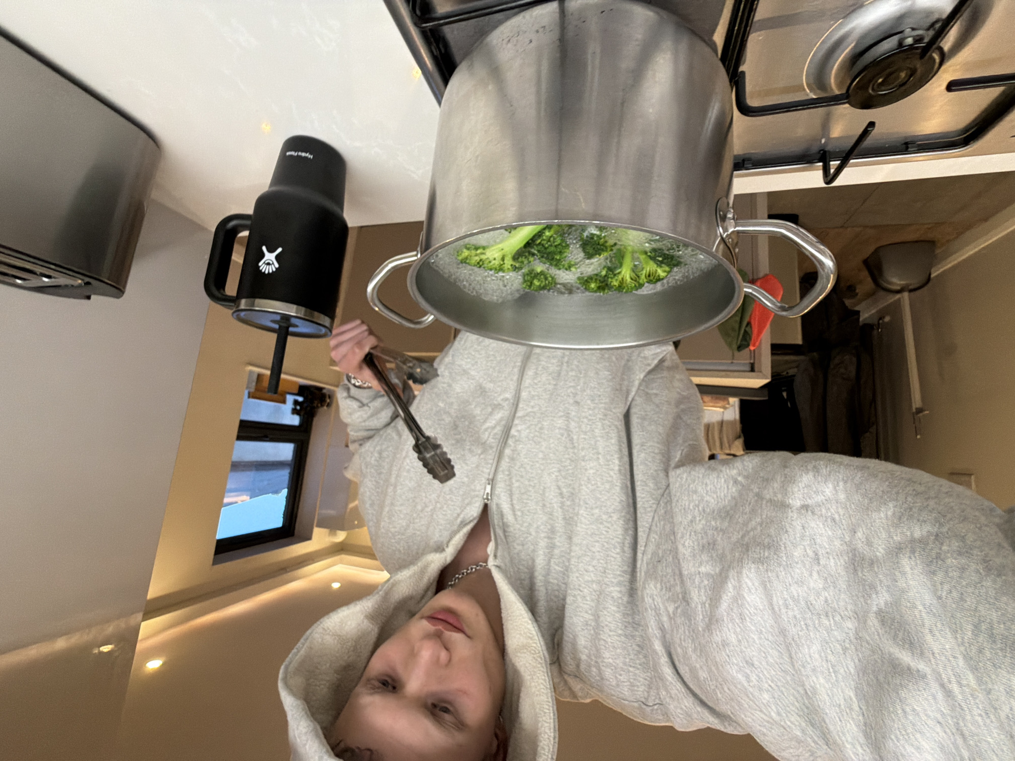This image captures a rotated, medium close-up view of a person cooking broccoli in a kitchen. The individual, appearing male with fair skin and light hair, is wearing a light grey zippered hooded sweatshirt and a silver chain necklace, and looks directly at the camera with a neutral expression. They hold a pair of metal tongs in their left hand.
The main activity shows a large, silver metal pot on a gas stovetop, where green broccoli florets are actively boiling in water, with visible steam and bubbles. To the left of the pot, on a white countertop, stands a black Hydro Flask coffee mug with a handle. The mug prominently displays the white Hydro Flask logo (a stylized 'X' within a circle), and the text "Hydro Flask" is visible on its lid.
The kitchen setting includes white countertops and what appears to be a gas stovetop with black grates. A silver-toned appliance is partially visible on the far left. In the background, through a window, a bright blue sky and some distant structures suggest it is daytime. Recessed lighting is visible on what would be the ceiling or under cabinets. The entire image is rotated approximately 90 degrees counter-clockwise, giving an inverted perspective of the scene. The location is specified as London, United Kingdom.