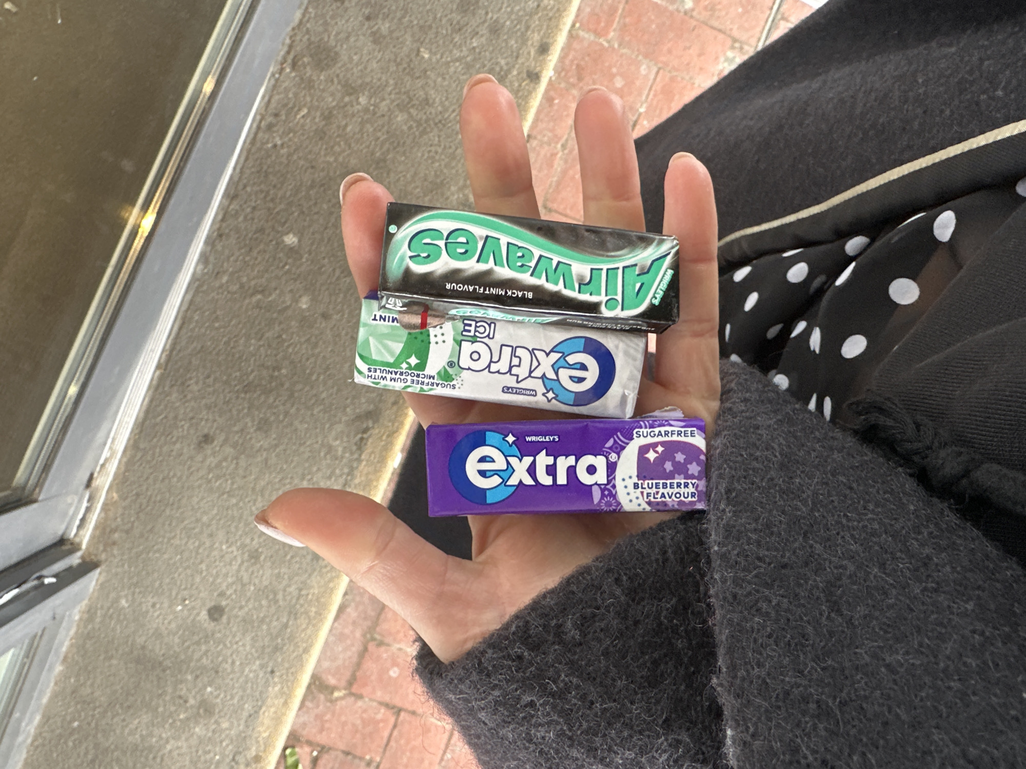 A human left hand is centrally positioned, displaying three packets of chewing gum. The hand exhibits short fingernails with a light, possibly nude-colored polish, showing minor wear at the tips. The gum packets, from left to right, are: a purple "Extra" brand packet, identified as "BLUEBERRY FLAVOUR" and "SUGARFREE"; a white and green "Extra ICE" packet, indicated as "MINT" and "SUGARFREE GUM WITH MICROGRANULES"; and a black and green "Airwaves" packet, labeled "BLACK MINT FLAVOUR" and "WRIGLEY'S".
The individual's dark grey, textured sleeve is visible on the left side of the frame, covering part of the forearm. Below the hand, a black garment with white polka dots is discernible, adjacent to a plain dark fabric segment featuring a vertical, thin, light-colored stripe.
The background consists of an urban outdoor setting. In the foreground, a rough grey concrete surface transitions to a section of red brick paving, laid in a stretcher bond pattern. Above this, a dark-framed window or door with reflective glass is visible, suggesting a building facade. The precise location within Tunbridge Wells, United Kingdom, cannot be determined from the available visual information, but the environment is consistent with a public street or pedestrian area.