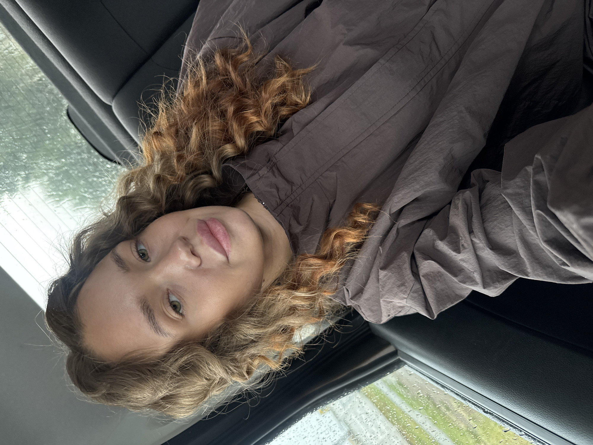 A young woman with curly, light brown hair and light green eyes is looking at the camera. She is wearing a brown jacket. She is sitting in the back seat of a car. The window beside her is wet with rain, and the view outside is blurry green and gray. The interior of the car is dark, with black leather seats visible. The image appears to have been taken in the city of Chaikovsky, Russia.