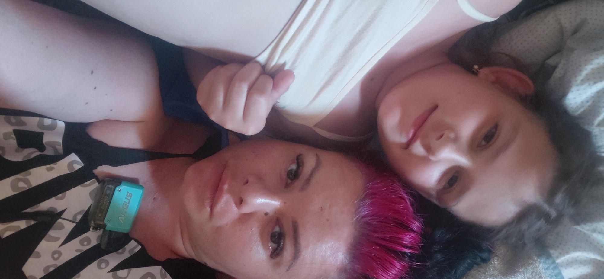Two people, likely an adult and a child, are lying down close together, looking up at the camera. They are in a horizontal orientation. The person in the foreground has bright pink hair, dark eyes, and is wearing a black top with a white patterned design. A turquoise object, possibly a vape or electronic device, is visible on their chest, with the word "ARGUS" printed on it. The person in the background has lighter skin and dark hair. They are wearing a light-colored top. The setting appears to be indoors, possibly a bed, given the soft bedding visible. The lighting suggests it might be daytime, but there are no strong indicators of the time of day or weather conditions. The overall mood seems relaxed and intimate.