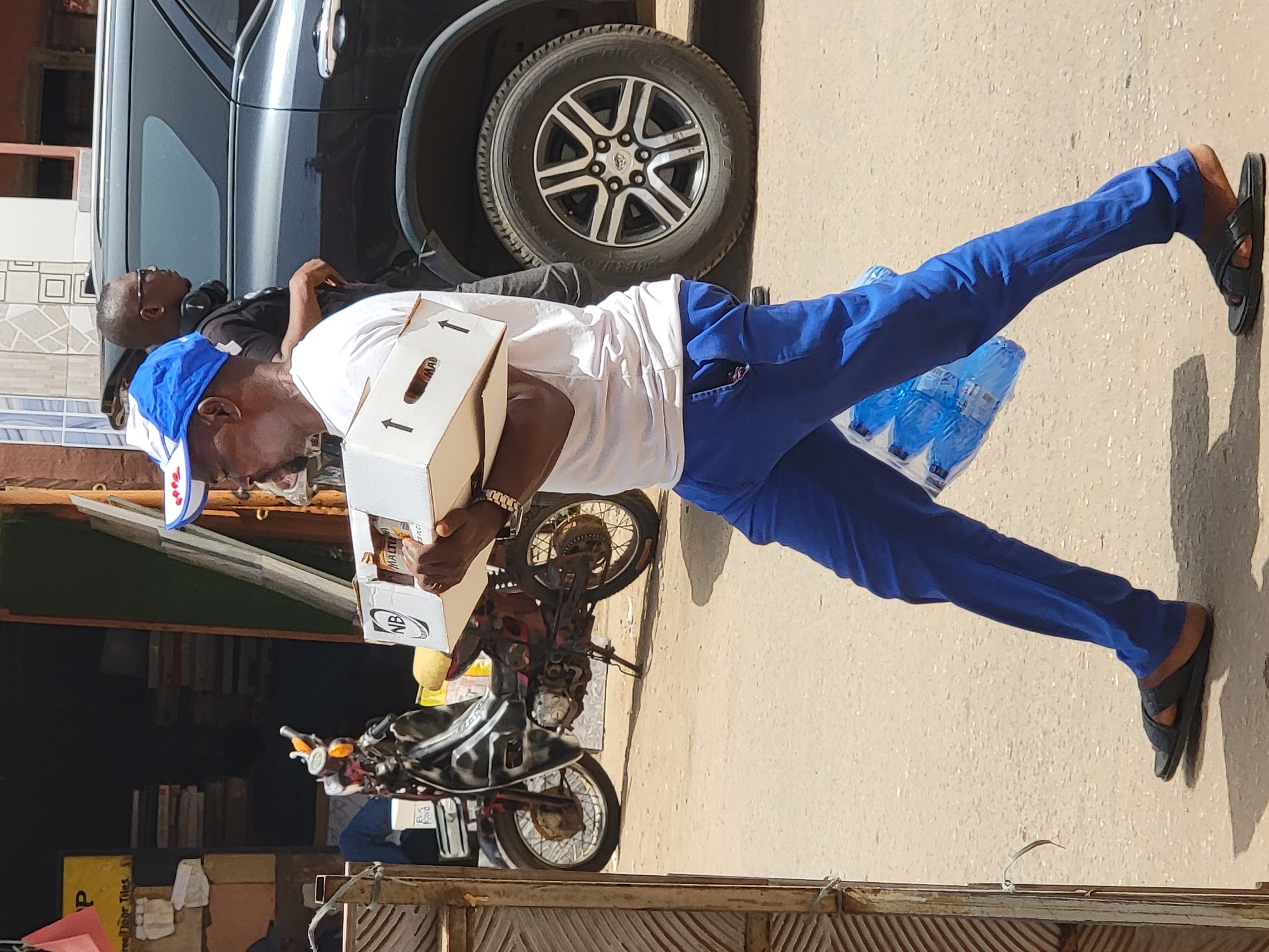 A man stands outdoors in Maiburiji, Nigeria. He wears a white t-shirt, blue pants, and sandals, and a blue cap with white writing. He carries a white box with "NB" printed on it, and a six-pack of bottled water. Another man is nearby, near a black SUV. A motorcycle is parked near the man. The ground appears to be paved or a dirt road.