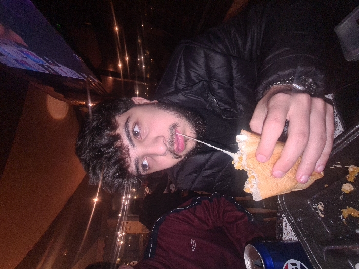 A man in a black puffer jacket eats a sandwich with stringy cheese, tongue out. He wears a silver watch on his left wrist. A dark metal tray beside him holds the sandwich. A partially visible beer can sits near the tray's edge. Soft lights are visible behind the man. He has dark hair and a beard. A television screen is visible behind him. Part of another person is visible wearing a red shirt.