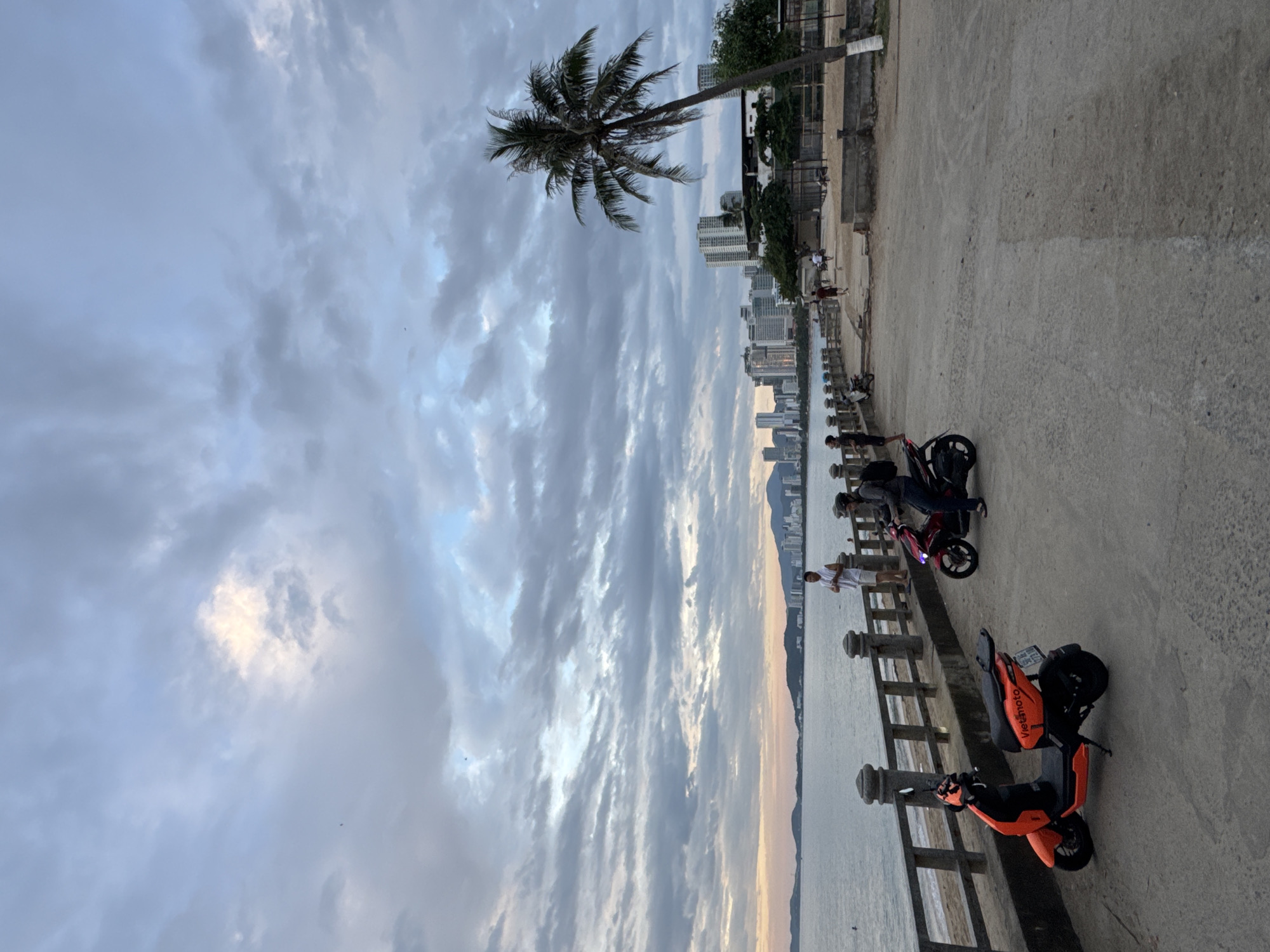 The image depicts a cloudy sunset over the ocean in Nha Trang, Vietnam. A promenade runs along the coast, with a railing separating it from the beach and the sea. Several people are present on the promenade, some standing and observing the view, while others are on or near motorbikes. Two motorbikes are prominently featured in the foreground: one is bright orange and parked, and the other is red and appears to be in use by a person wearing a helmet. In the background, a cityscape with numerous tall buildings lines the coast, and a lone palm tree stands on the right side of the frame, silhouetted against the sky. The overall atmosphere is calm and suggests a public gathering space enjoyed during the evening.