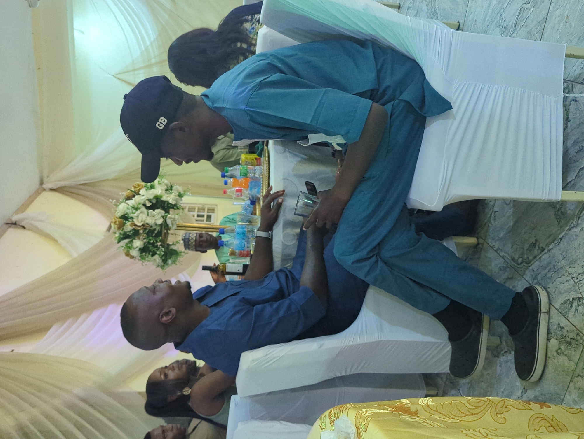 Two men in matching blue traditional attire are seated at a table during a gathering in Barden Yana, Nigeria. The man on the left is facing the man on the right, with his mouth open as if speaking. He is holding a clear glass in his right hand. The man on the right is wearing a navy blue baseball cap with "GB" on it and is looking towards the man on the left. The table is covered with a white tablecloth and has several bottles of beverages and a floral centerpiece. In the background, other guests are visible, and the area is decorated with draped white fabric, suggesting an indoor event.