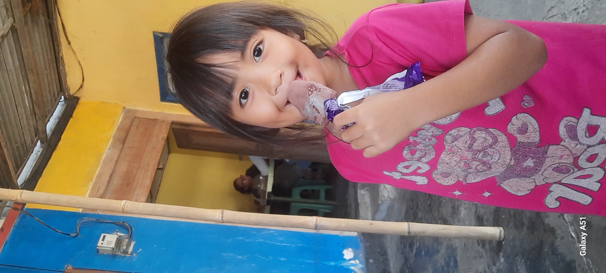 A young girl with dark hair and bangs smiles at the camera while holding a purple-wrapped chocolate ice cream bar to her mouth. She is wearing a bright pink t-shirt with a graphic of a teddy bear wearing glasses and the word "BOY" printed on it in large letters, with smaller hearts scattered around the design. She stands in front of a yellow wall and a blue wall, with a bamboo pole to her left. In the background, through an opening, another person is visible seated at a table in what appears to be an indoor setting with a yellow wall. The floor is a gray, rough surface. The image also includes a power meter mounted on the blue wall. The bottom right corner of the image shows "Galaxy A51".