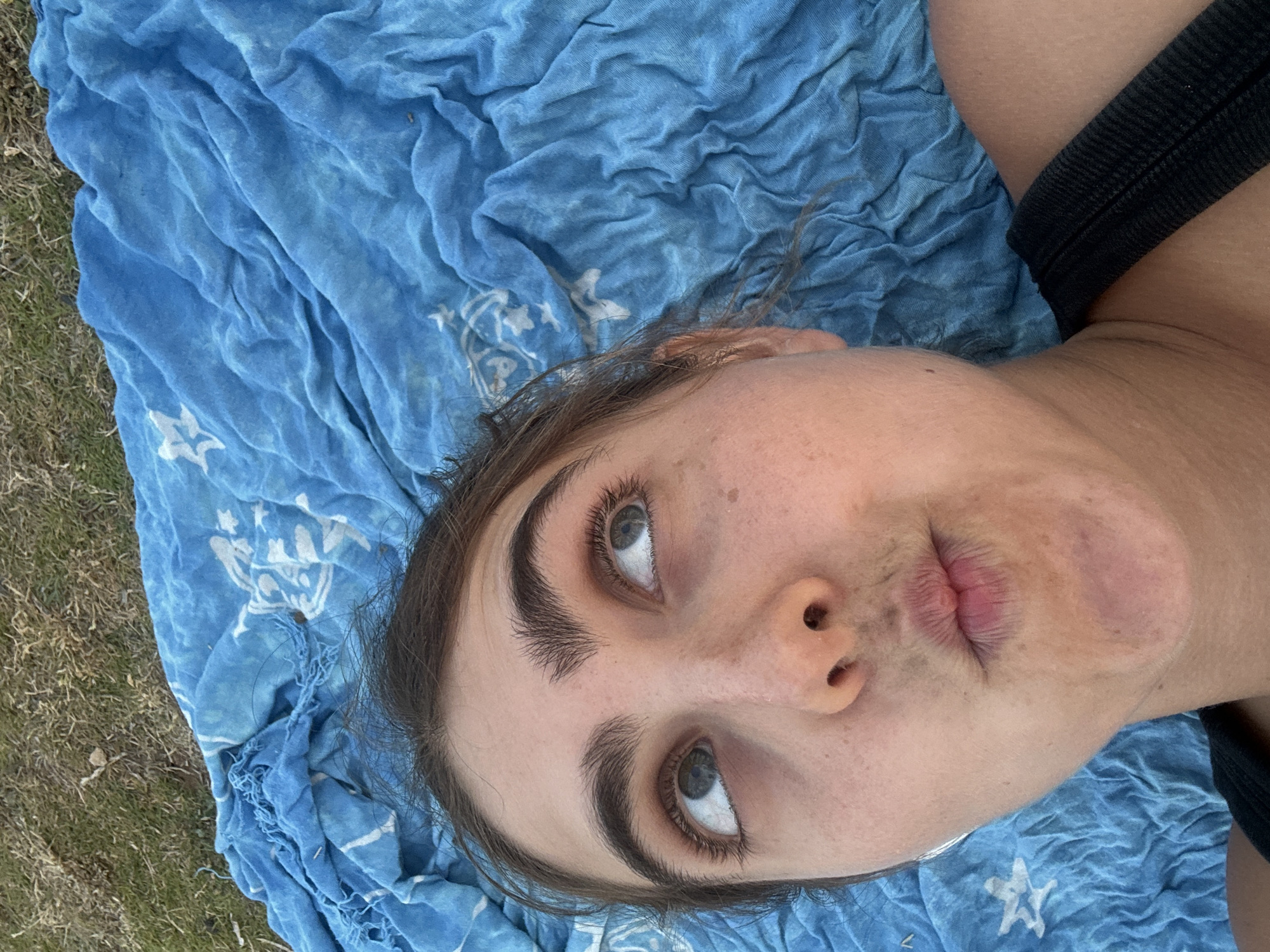 A young woman is lying on a blue towel on grass, looking up and to the right with a pouty expression. She has dark eyebrows and eyelashes, and her eyes are visible. The towel is draped behind her head and shoulders, and appears to be made of a lightweight fabric with a pattern of white stars. The background is out of focus, showing grass and possibly other foliage. The woman is wearing a black strap, possibly from a tank top. The lighting suggests an outdoor setting during the day.