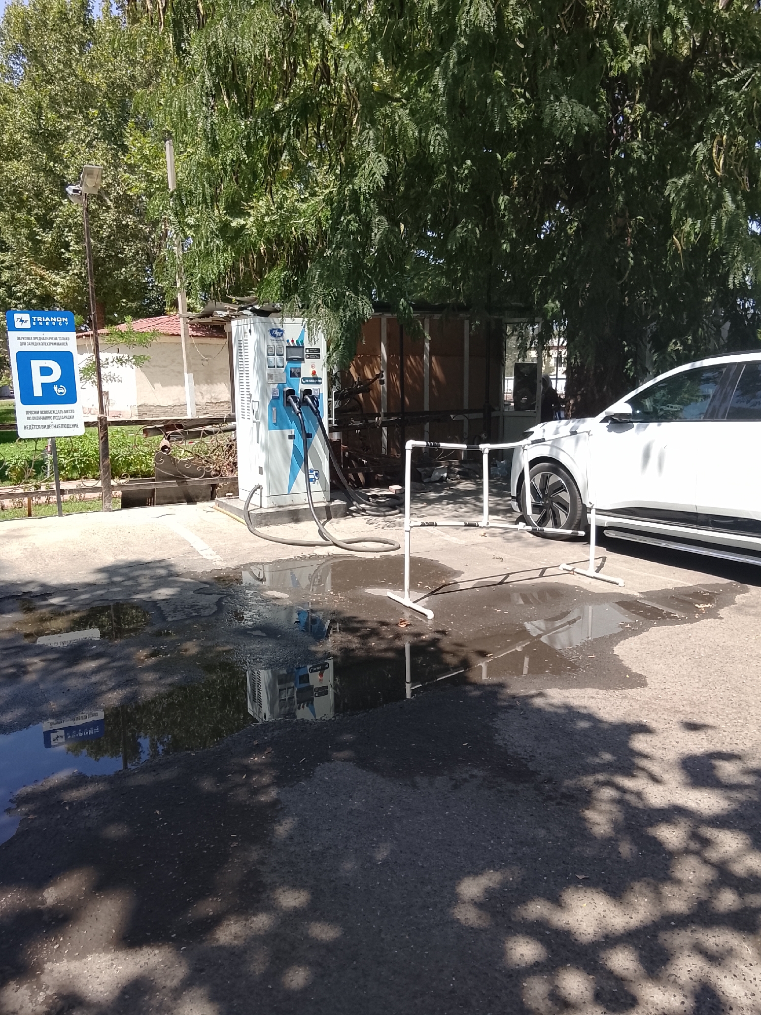 The image depicts an outdoor scene in Fergana City, Uzbekistan, featuring an electric vehicle charging station. A white electric car is parked next to a tall, white charging station with blue accents. Two charging cables extend from the station, one of which is connected to the car. A blue and white parking sign with Cyrillic text is visible to the left, indicating the area is reserved for charging electric vehicles. The ground is wet with puddles, suggesting recent rain or a spill. In the background, trees provide shade, and a building structure is partially visible. The overall environment appears to be a public parking area or lot.