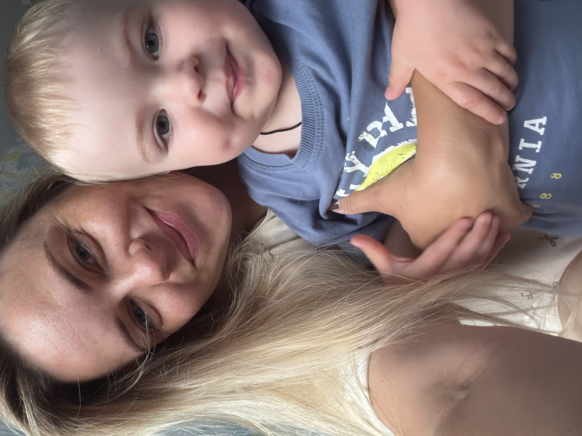 This is a close-up, selfie-style image of a woman and a young child. The woman, with blonde hair and fair skin, is looking towards the camera with a slight smile. Her face is in focus in the foreground. The child, with blonde hair and blue eyes, is positioned behind and slightly above the woman, with their head resting on her shoulder. The child is looking slightly away from the camera.
Both subjects are wearing casual clothing. The child is wearing a blue t-shirt with yellow lettering on the front and a thin black cord around their neck. The woman is wearing a light-colored top, possibly a t-shirt or tank top. Their hands are intertwined, with the child's hand resting on the woman's arm.
The overall mood of the image appears warm and affectionate, suggesting a close bond between the two. The lighting is soft, indicating it could be daytime, though no specific time is discernible. There are no clear environmental cues to suggest the specific location within Votkinsk, Russia, or the weather conditions.
The visible text on the child's shirt appears to be part of a word, possibly "NIA" is visible, along with some smaller yellow markings that resemble numbers or symbols. The image is oriented vertically.