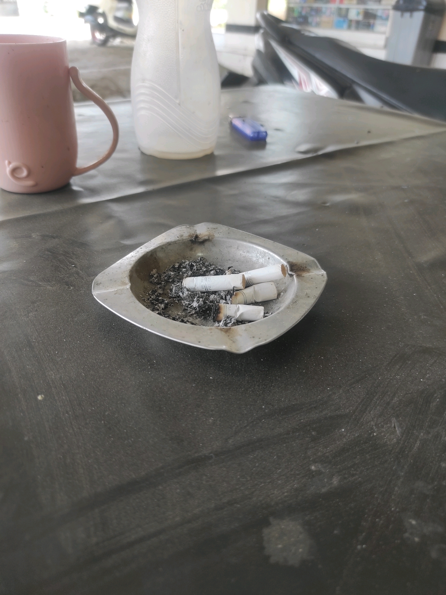 The image depicts a metal ashtray filled with cigarette butts and ash, placed on a dark, textured surface. To the left of the ashtray is a pink mug with a curved handle. In the background, a white plastic bottle or container is visible, and further back, a portion of a motorcycle seat and handlebars can be seen. A blue lighter rests on the surface near the motorcycle. The overall environment appears to be outdoors or in an open-air setting, possibly a street or a shaded area in Cipada, Indonesia, given the presence of a motorcycle and the typical lighting conditions. The scene suggests a moment of pause or rest, with evidence of smoking.