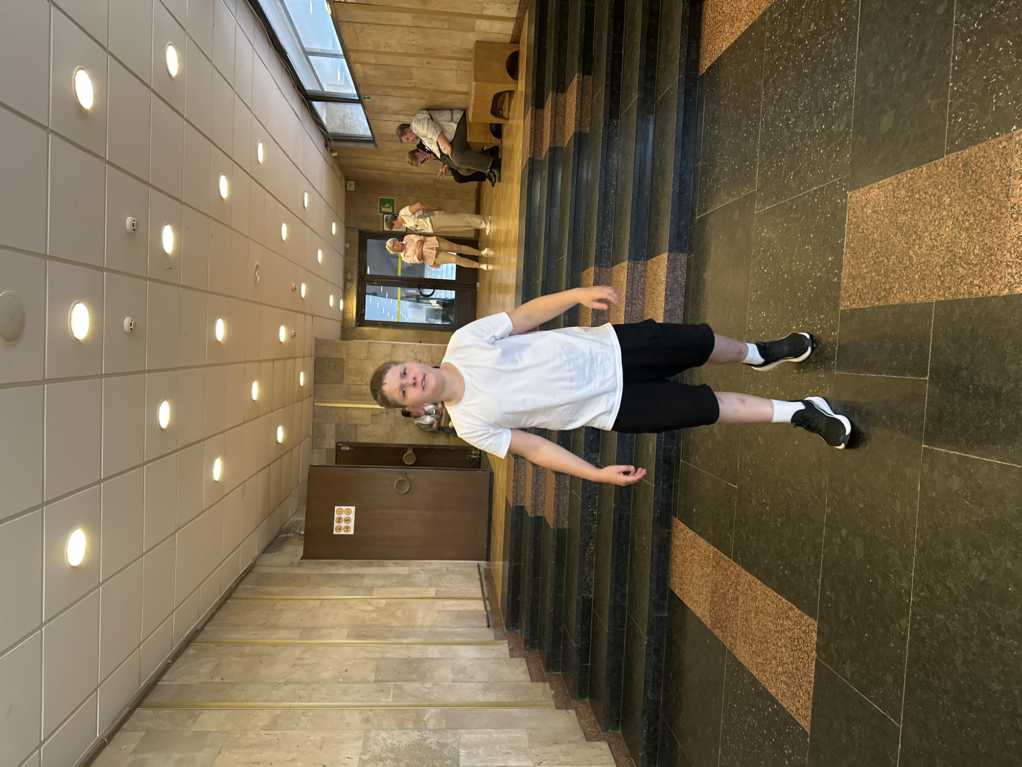 The image depicts an indoor public space, specifically a multi-level corridor or lobby within a building in the city of Unknown, Unknown. The scene is illuminated by numerous circular recessed LED lights embedded in a white grid-panel ceiling, supplemented by natural light from horizontal windows on the far right wall.
The flooring consists of large, dark gray/black polished stone tiles with light speckling, interspersed with rectangular sections of brown-orange speckled granite. A series of wide steps, constructed from the same dark gray/black polished stone with brown-orange granite risers, creates a two-tiered floor plan. The walls are clad in light-colored, vertically oriented stone tiles, possibly marble or travertine.
In the foreground, slightly left of center, a young male is walking towards the viewer. He wears a plain white short-sleeved t-shirt, black cargo shorts, white crew socks, and black athletic shoes with white soles. His expression is neutral.
In the midground, to the left, a large dark brown wooden door with a round metallic handle is visible at the top of the steps. Above this door, a white sign displays six yellow, emoji-like circular symbols. To the right of the steps, further back, a set of double glass doors leads to another interior space. Above these glass doors, a green "exit" sign is affixed.
Multiple individuals are present in the background. Near the glass doors, two women are standing and conversing: one in a light pink short-sleeved shirt and beige pants, the other in a white t-shirt and light-colored pants. To their right, two men are seated on a light brown upholstered bench with wooden legs. One man, wearing a striped shirt and dark pants, is looking down at a mobile phone. The other man, dressed in a light-colored polo shirt and dark pants, is looking generally forward. Faintly visible behind the dark brown door on the left, another person is seated.
The ceiling features two small, white, circular smoke detectors. A horizontal ventilation grille is present high on the left wall.