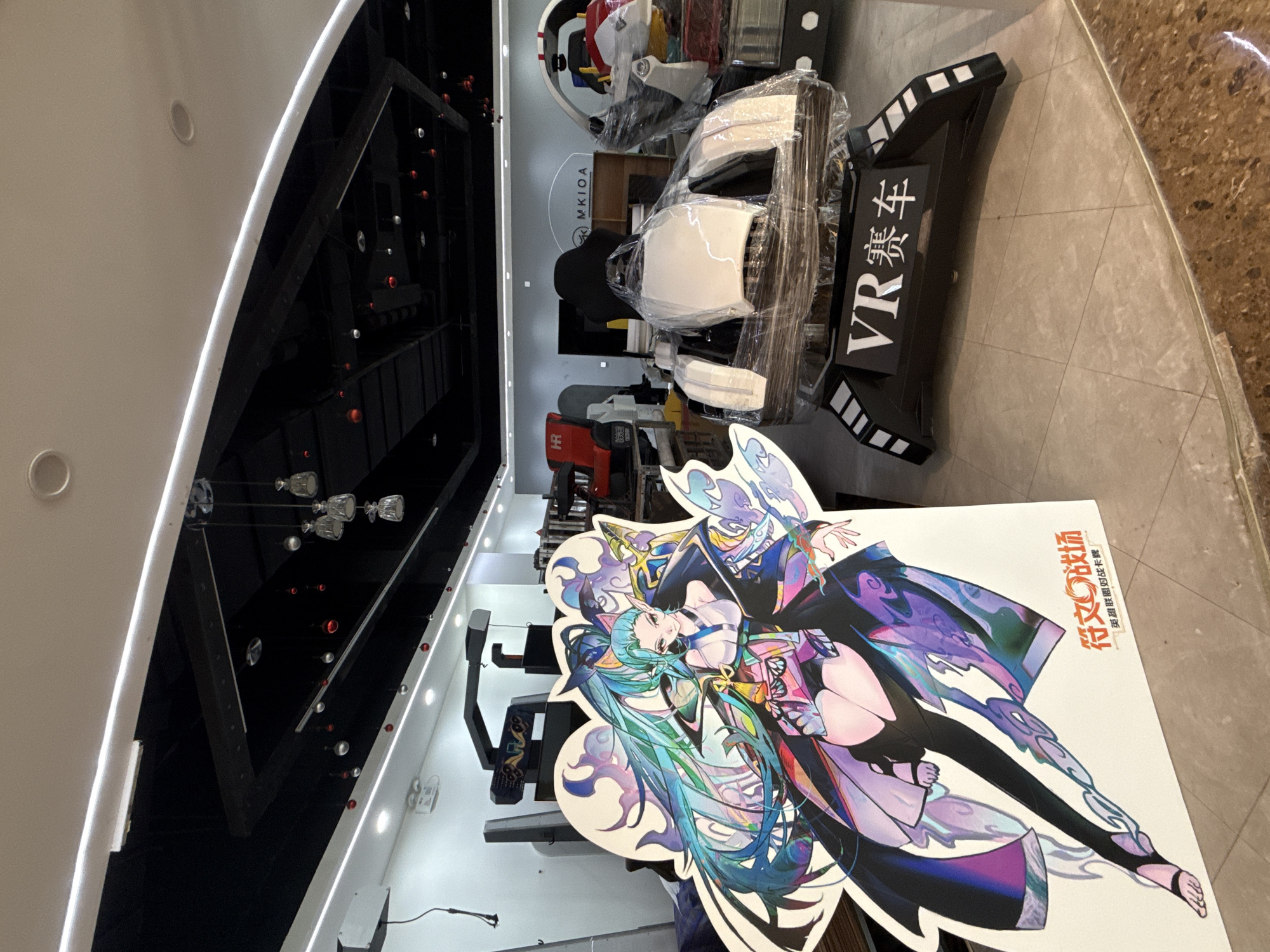 In a retail or exhibition space in Huizhou, China, a large cardboard cutout of an anime-style female character with long, flowing teal and purple hair stands prominently in the foreground. To the right of the cutout is a futuristic-looking VR racing simulator, partially wrapped in clear plastic. The simulator has a white seat and a black base with the Chinese characters "VR 赛车" (VR Racing Car) visible. In the background, various other arcade machines and display units are visible, some with red accents. The ceiling features a black structure with embedded lights and hanging decorative elements, possibly Christmas ornaments. The floor is tiled, transitioning to a wood-like surface in the background. The overall impression is that of an entertainment or gaming venue, potentially preparing for an event or a new display.
