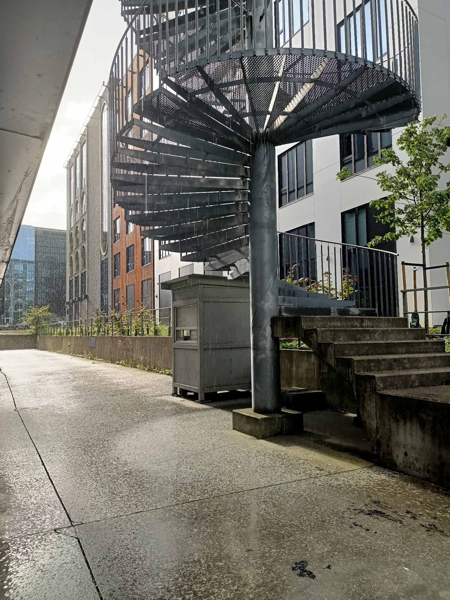 The image depicts an outdoor scene in Tallinn, Estonia, characterized by wet pavement and a light rain. A prominent spiral staircase made of metal occupies the right side of the frame, leading up to a walkway. Adjacent to the staircase are concrete steps. In the background, modern buildings with large windows are visible. A small, grey, booth-like structure stands near the base of the spiral staircase. Greenery, including trees and bushes, is present throughout the scene. The ground appears to be a paved area, possibly a courtyard or walkway, with visible cracks and water puddles. The overall atmosphere is overcast and damp, suggesting inclement weather.