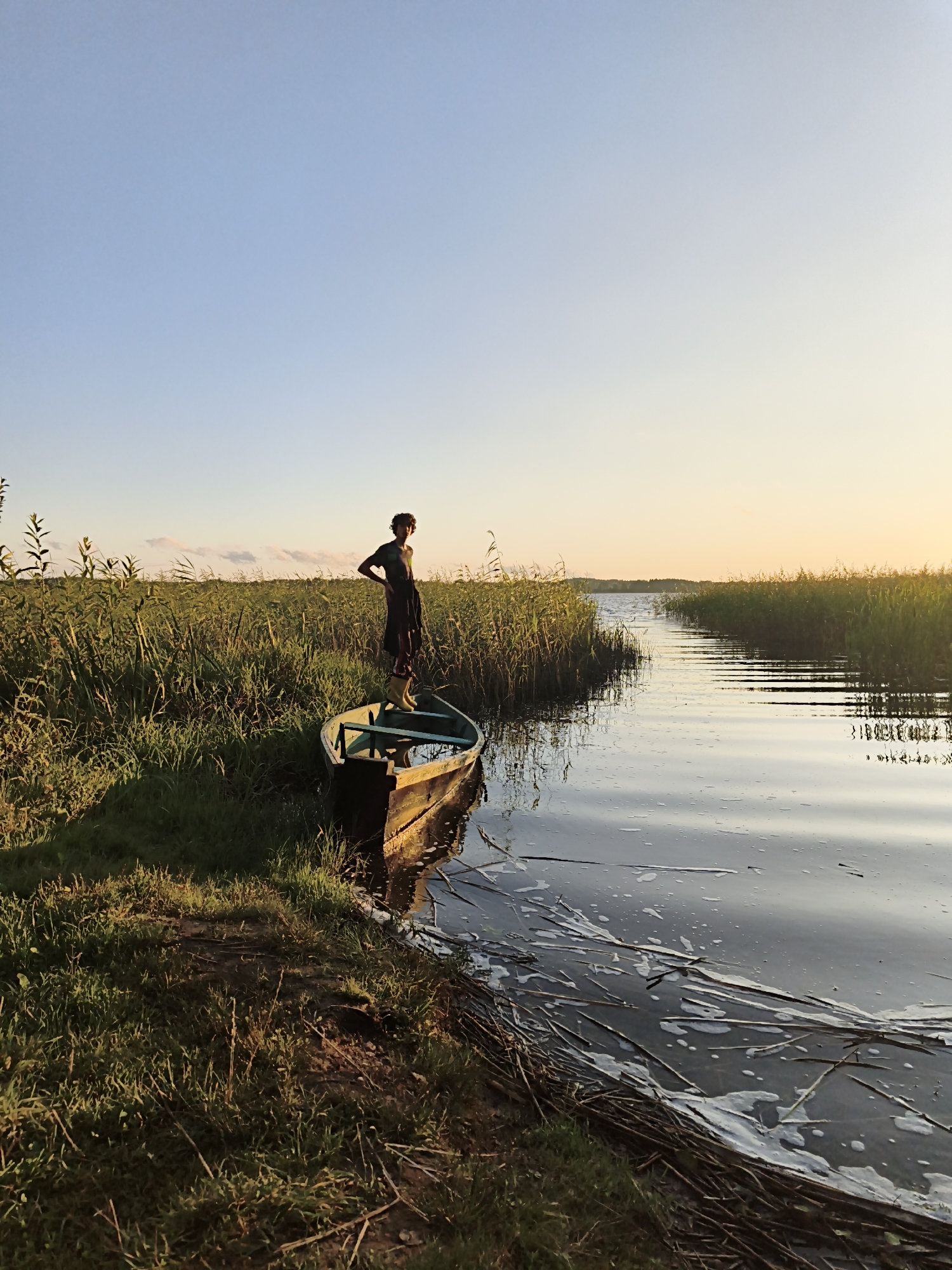 In the vicinity of Свірскі сельскі Савет, Belarus, a person stands in a small wooden boat anchored near a grassy bank. The boat, weathered and filled with water, is partially submerged. The person, dressed in casual clothing and rubber boots, stands with one hand on their hip, facing away from the camera towards a body of water. The water is calm, reflecting the pale blue sky and the warm glow of the setting sun. Tall reeds and grasses line the water's edge, creating a dense natural barrier. The overall scene depicts a tranquil, natural environment at dusk.