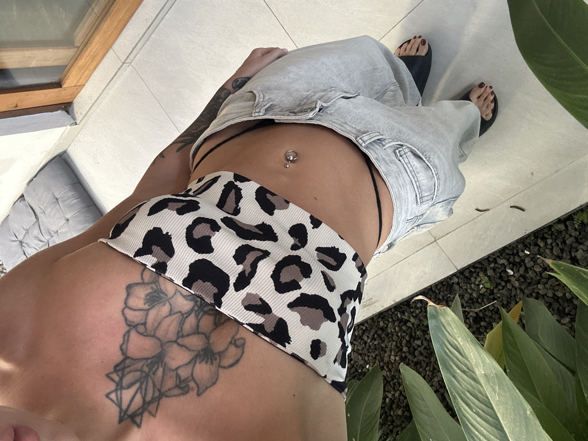 A woman's torso and legs are visible in a partially obscured overhead shot taken outdoors. She is wearing a bikini bottom with a leopard print design and low-rise, light wash denim pants. A navel piercing is visible. She has tattoos on her torso and arm. The woman's feet are clad in dark-colored sandals. The setting includes light-colored tiled flooring, greenery, and a grey cushioned piece of furniture. Based on the request's reference to Dalung, Indonesia, the location is assumed to be there.