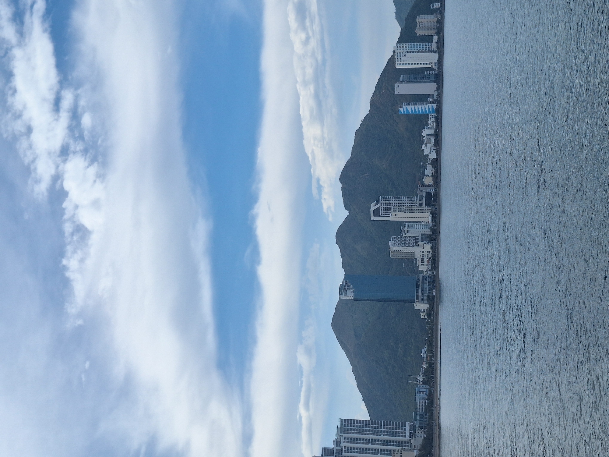 A panoramic view shows the coastline of Nha Trang, Vietnam, featuring a body of water in the foreground and a city skyline backed by lush green mountains under a cloudy sky. The water is a deep blue with subtle ripples. The city skyline is composed of numerous modern high-rise buildings of varying heights and designs, predominantly white and grey, with a few featuring blue facades. A distinct tall, slender building with a blue exterior stands out amongst the others. Behind the city, a large, mountainous landmass covered in dense green vegetation dominates the background, its peaks softened by distance and atmospheric haze. The sky is a mix of blue and white, with streaky clouds creating a dynamic pattern. There are no discernible people or actions depicted in the image.