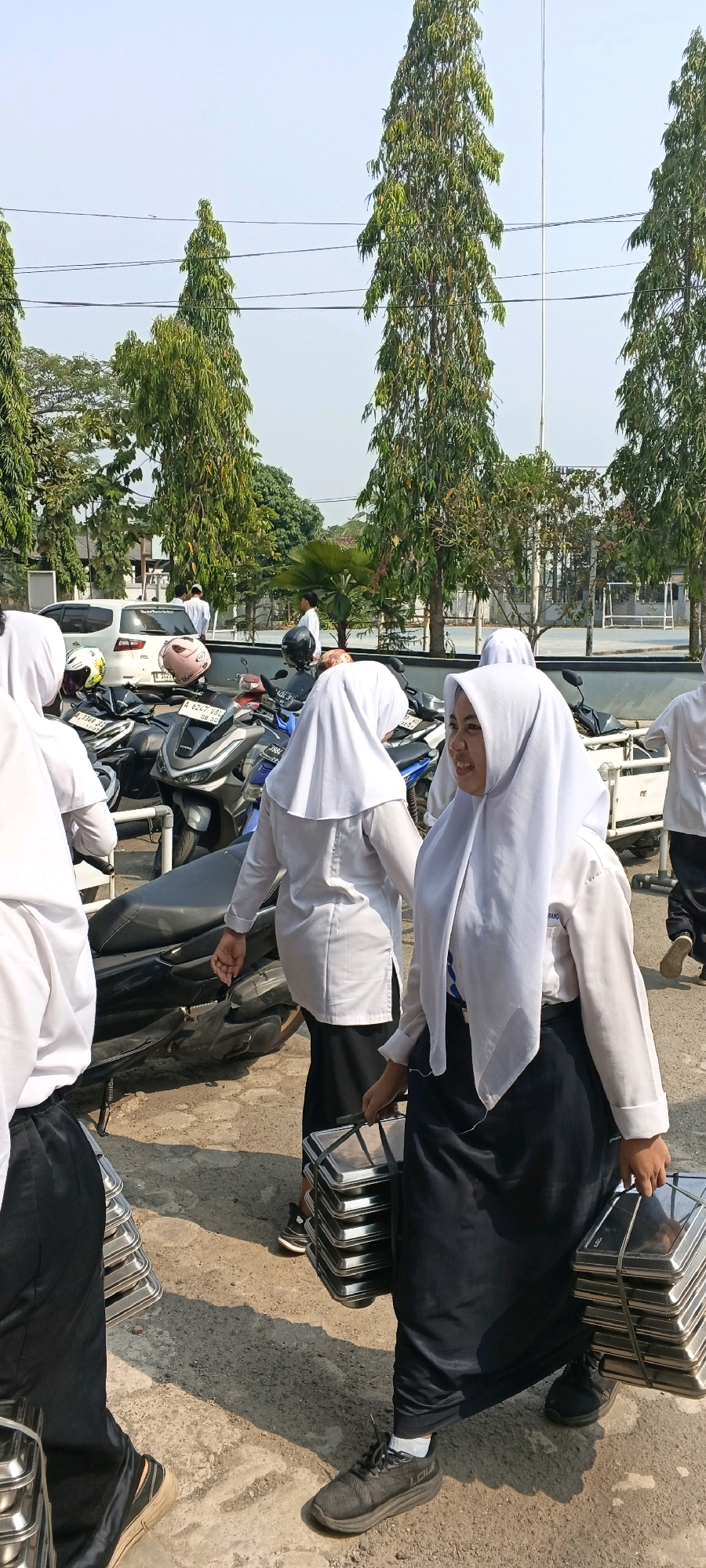 In Suka Tani, Indonesia, a group of individuals, predominantly young women wearing white hijabs and dark skirts or pants, are captured in an outdoor setting, likely near a school or community gathering. The environment suggests a sunny day with a clear sky, bordered by tall, slender trees. In the background, a white car and numerous motorcycles are parked, indicating a common mode of transportation in the area. The central focus is on the individuals carrying stacks of metal trays, which appear to be used for serving food. One woman in the foreground, smiling and looking to her right, holds a stack of trays with both hands, her face illuminated by the sunlight. Other individuals are also seen carrying similar trays, suggesting a collective effort in food distribution or preparation. The ground is paved with a rough, sandy surface, common in many outdoor Indonesian locales. The overall scene depicts a practical, communal activity taking place in a typical Indonesian urban or suburban environment.