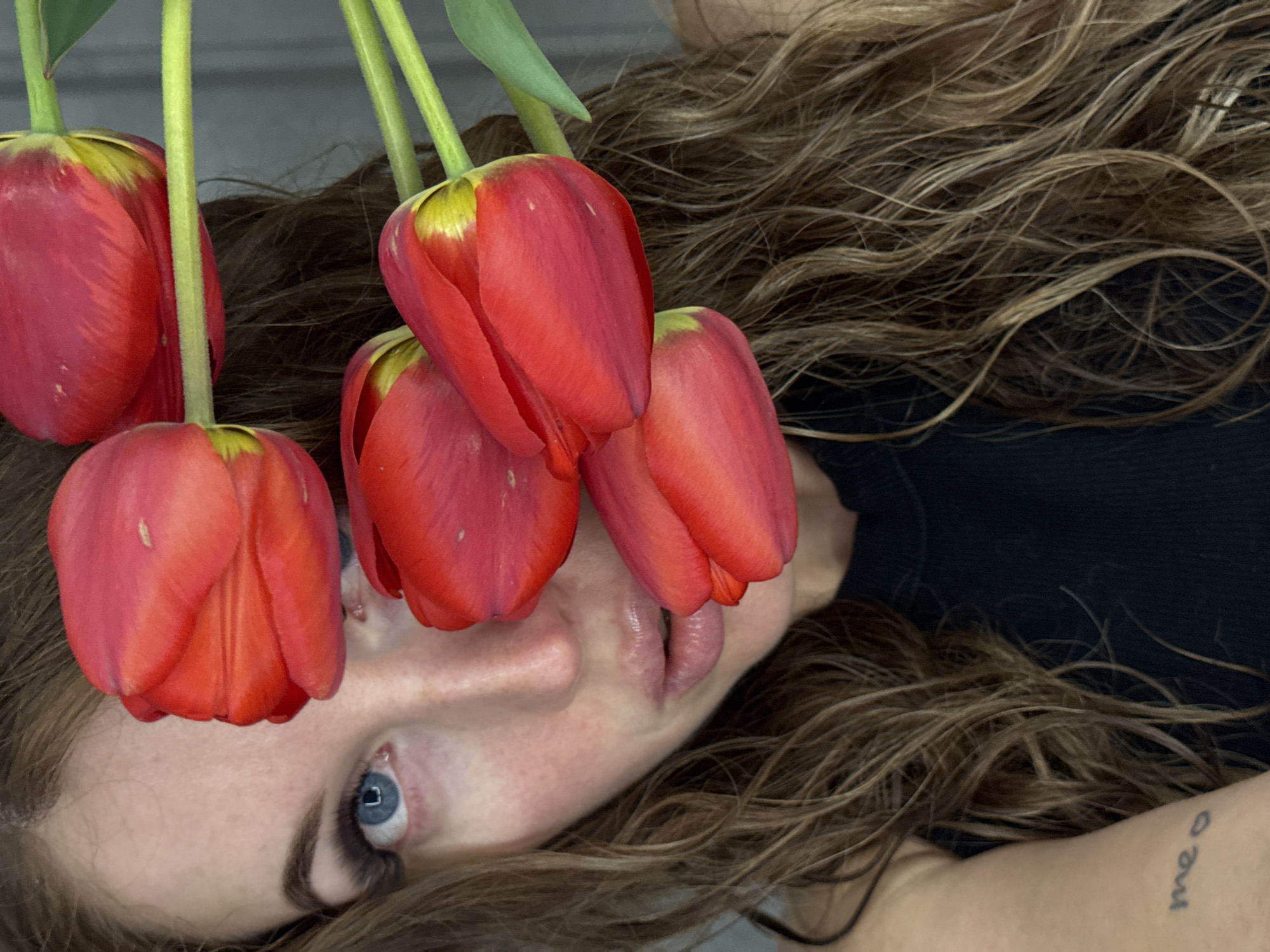 The image shows a woman lying down with red tulips positioned over her face. The woman has fair skin, brown hair, and blue eyes. Her lips are visible beneath the flowers. A tattoo is visible on her arm. The background is blurry and appears to be a neutral color. The image does not contain information to determine its location within Chaikovsky, Russia.
