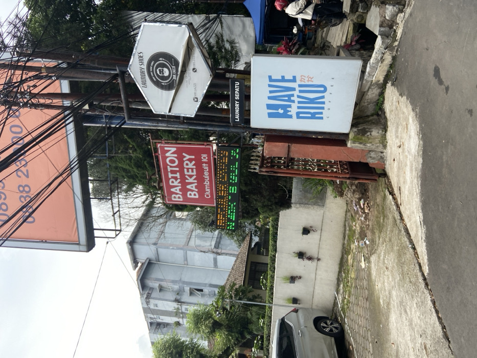 The scene is located in Bandung, Indonesia, at coordinates -6.88 latitude and 107.603 longitude. It shows a street with a gray car parked on the left, facing a white wall with a gate. On the right side of the image, a black pole is attached to several signs. The top sign is white with a circular red logo for "Laundry Shoes" and the text "The Lazy." Below it is a red sign that reads "Bariton Bakery Clumbuleuit 101." Below this is a green digital sign displaying a price list. The bottom sign is blue and white and reads "Mave Riku Coffee." There is a blue canopy in the background and a person in a red jacket standing near a motorcycle. The environment includes a concrete parking lot, a wall with a metal gate, and a sidewalk in front of a coffee shop. There is debris scattered on the ground.