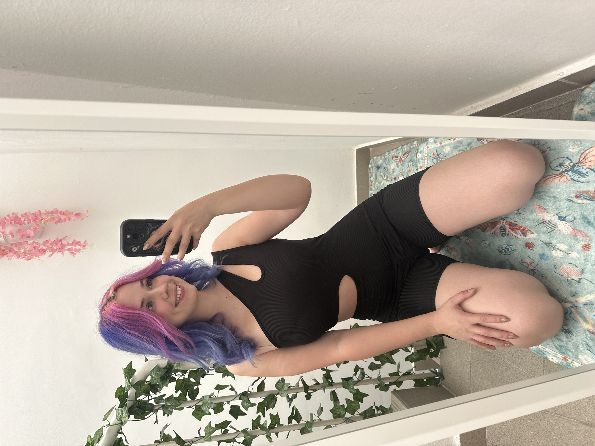 A young woman is captured in a mirror selfie, smiling brightly at her smartphone. Her hair is distinctly dyed with vibrant pink on one side and blue-purple on the other. She is wearing a black, ribbed, sleeveless jumpsuit or romper that features two circular cutouts on her torso. She holds a black smartphone with a textured case in her left hand, positioned to take the photo, while her right hand rests gently on her bent left knee.
The reflection reveals an indoor setting. To the left, behind her, artificial pink flowers are arranged against a white wall. Below, a garland of artificial green ivy is draped over a white structure, possibly a railing. The floor beneath her is partially covered by a light blue rug adorned with patterns of insects and birds, and partially by light grey tiles. The upper part of the reflection shows a white ceiling and plain white walls, suggesting a brightly lit interior, likely during the daytime. The overall impression is one of cheerfulness and personal expression within a decorated domestic space. No visible text is legible in the image.