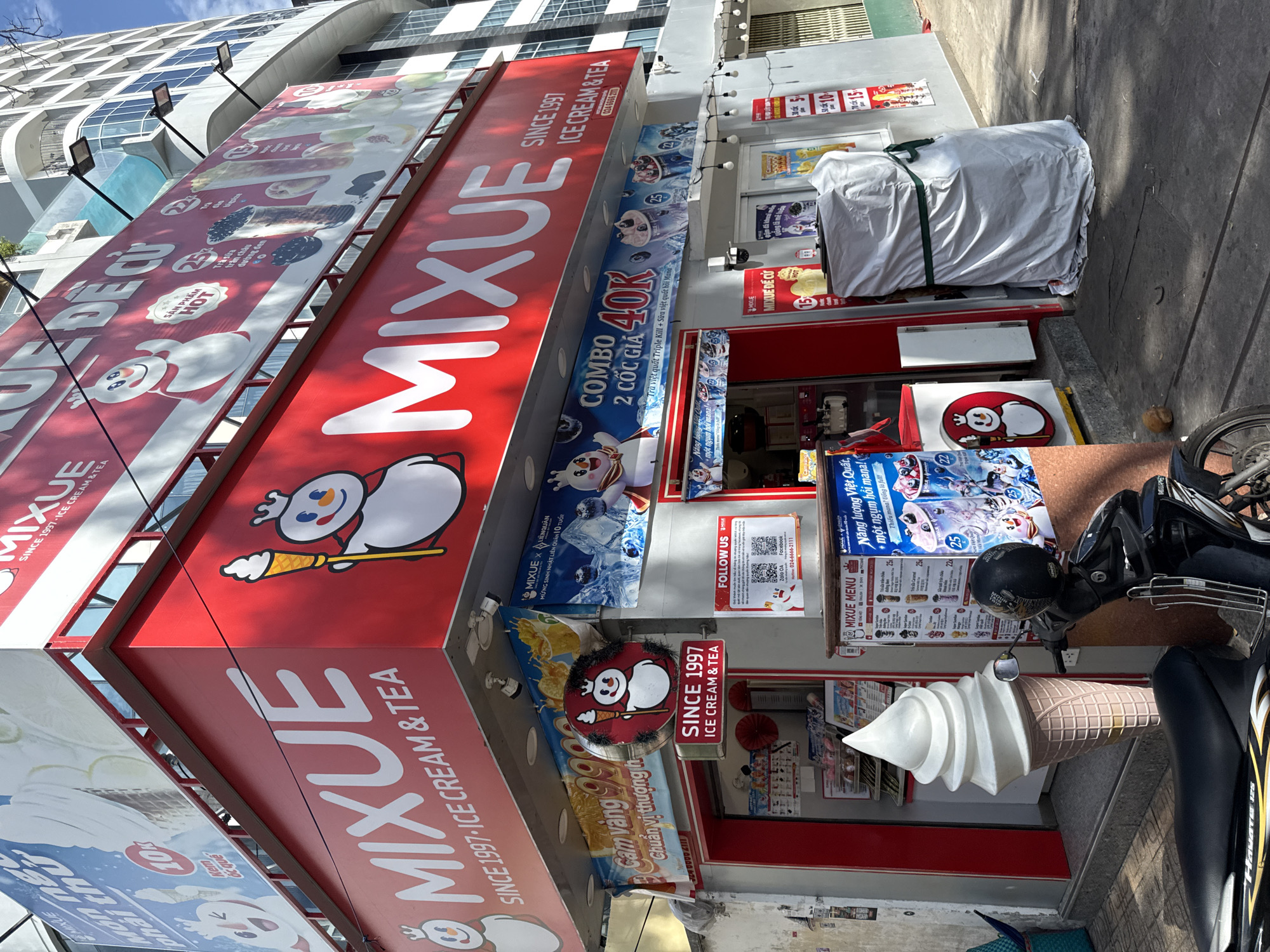 This image shows the exterior of a Mixue ice cream and tea shop in Nha Trang, Vietnam. The shop is brightly lit with red and white signage featuring the Mixue logo and mascot, a snowman character holding an ice cream cone. A large, white soft-serve ice cream cone sculpture is prominently displayed outside the shop. A black motorcycle is parked in front of the entrance. Various promotional posters and menus are visible on the shop's facade, advertising drinks and ice cream combinations. The building appears to be part of a commercial area with other businesses and a modern, multi-story building in the background. The overall scene depicts a typical street-level storefront in a Vietnamese city.
