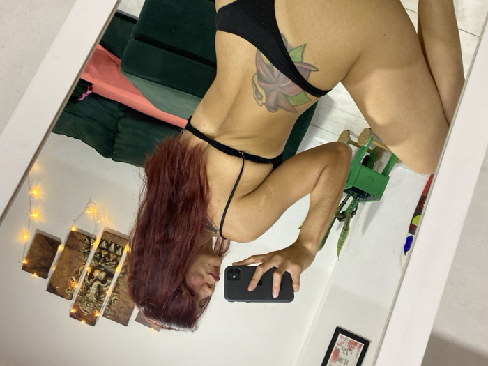 The image displays a woman taking a mirror selfie indoors in Wilde, Argentina. The woman, with long reddish-brown hair, is reflected in a white-framed mirror. She is wearing a black two-piece garment, possibly a bikini or underwear. A multi-colored flower tattoo is visible on her lower back/left hip. Her right hand holds a dark-colored smartphone, directed towards her reflection, with her lips slightly puckered. The background, reflected in the mirror, reveals a white wall adorned with various decorative elements: a framed picture with floral motifs, a multi-panel artwork depicting Ganesha, and a string of warm fairy lights draped over the Ganesha artwork. A dark green sofa or sectional, partly covered by a pink or red fabric, is also visible. To the left, a snake plant in a dark pot sits on a small green wooden stand, with another colorful object partially seen beside it. The overall environment suggests a residential interior.