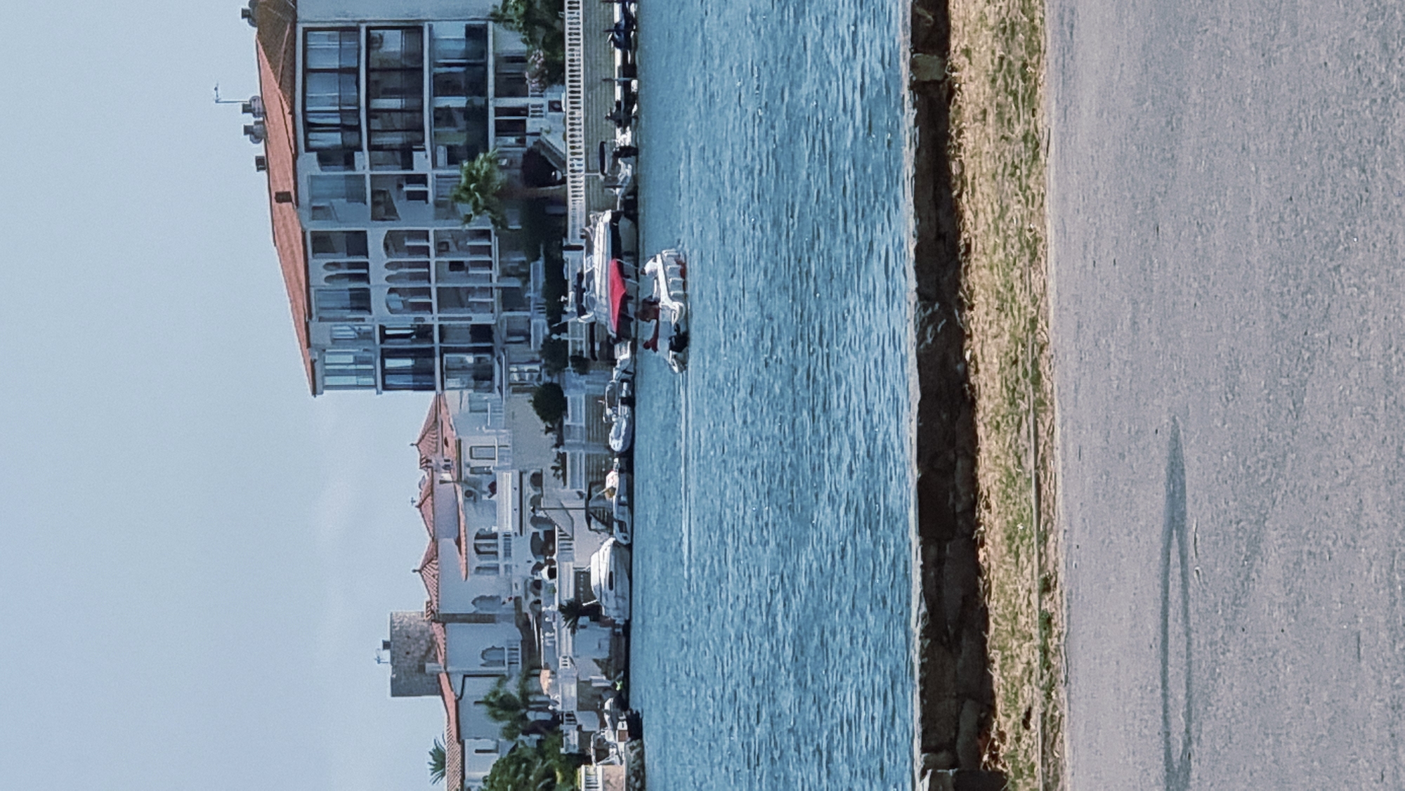 The image captures a serene waterfront scene in the city of Castelló d'Empúries, Spain, likely within the Empuriabrava marina area. In the foreground, a paved walkway or road runs horizontally across the bottom of the frame, with a stone wall and a strip of dry grass separating it from the water. The body of water, a vibrant blue, occupies the majority of the image.
On the water, a small, white motorboat with a red canopy is prominently featured, carrying two individuals. The boat is moving away from the viewer, leaving a faint wake behind it. Several other boats are docked or anchored along the shoreline in the mid-ground, including larger yachts and smaller vessels.
The background is dominated by a row of white buildings with red-tiled roofs, characteristic of Mediterranean architecture. These structures include a mix of multi-story apartment buildings with numerous balconies and windows, as well as more traditional houses. One notable building on the left appears to be a historic tower. Greenery, including palm trees, is visible interspersed among the buildings. The sky is a pale blue with a few wispy clouds. The overall impression is one of a sunny, tranquil day in a coastal town.