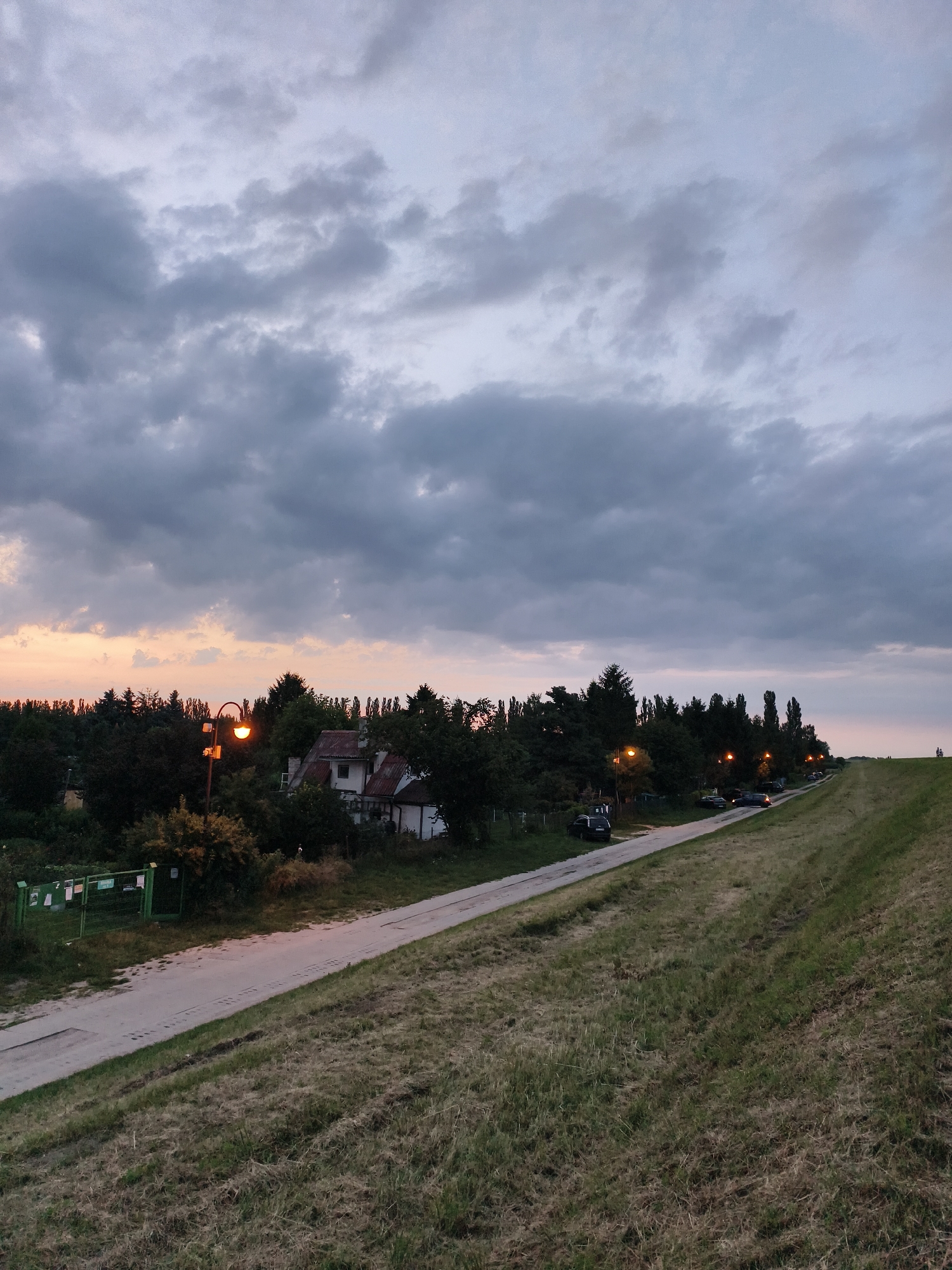 An eye-level, medium shot captures a view of a road lined with trees and streetlights in Tczew, Poland, under a cloudy twilight sky. The road curves gently to the right, with cars parked along its left side. A grassy embankment slopes upwards on the right side of the road. In the background, a cluster of houses with red-tiled roofs is nestled amongst the trees. The sky is filled with dramatic, grey clouds, with hints of orange and pink visible near the horizon, suggesting the sun is setting or has just set. The overall atmosphere is serene and peaceful.