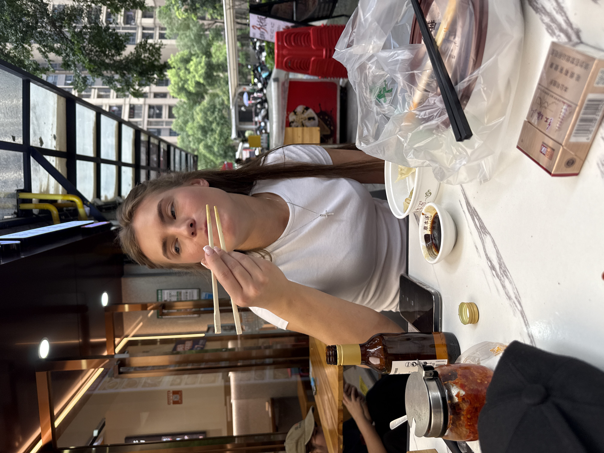 A young Caucasian woman with long brown hair is eating with chopsticks at an outdoor restaurant table in Haizhu District, China. She is wearing a white t-shirt and a silver cross necklace. She is looking to her right. In front of her is a white bowl of food, a small dish of soy sauce, and a bottle of beer. To her left is a jar of chili paste and a black baseball cap. In the background, there are buildings and trees, as well as other tables and chairs. The scene appears to be set in the afternoon, with natural light illuminating the area.