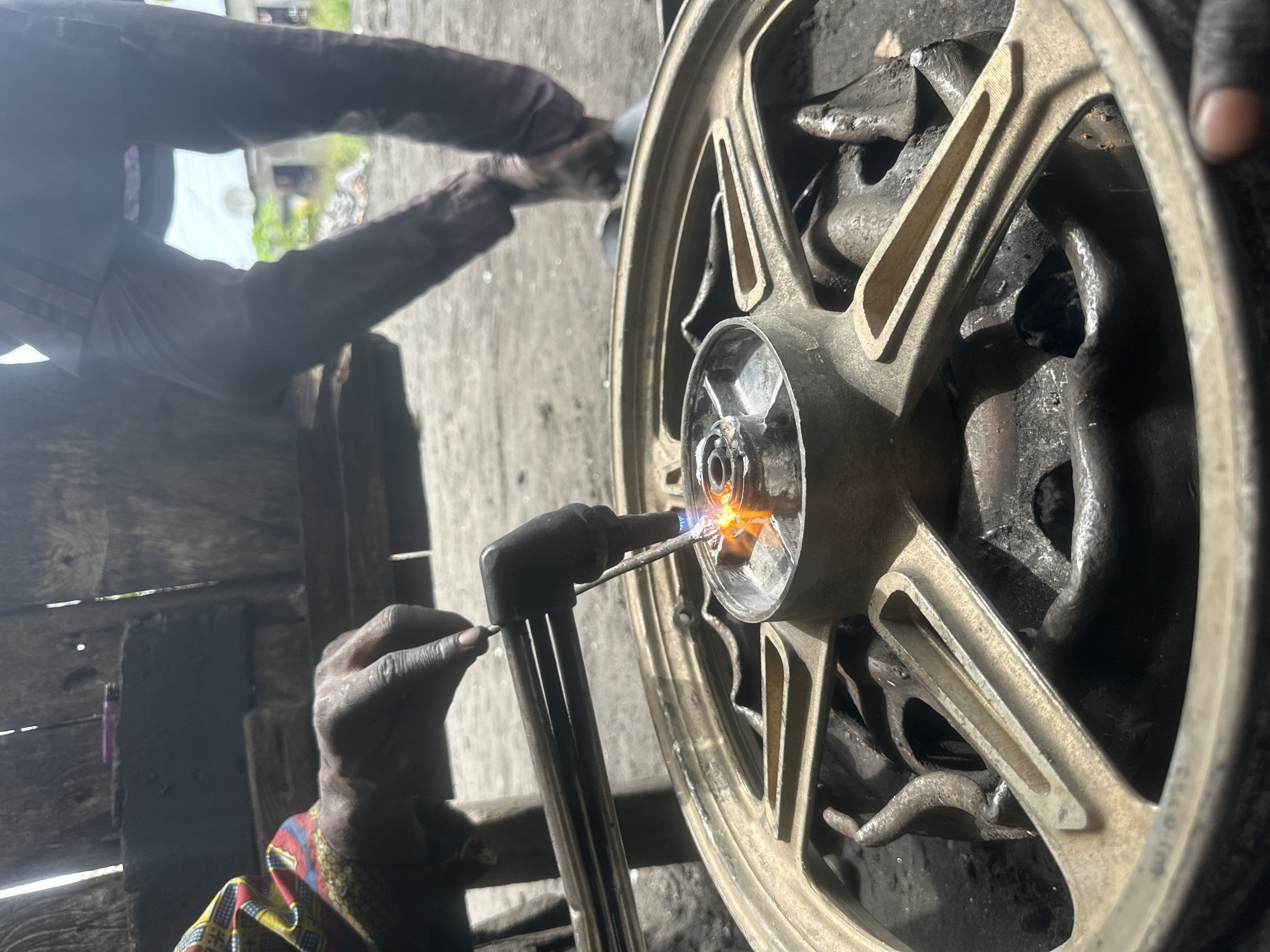 In this medium shot, two individuals are seen in what appears to be a workshop in Badagry, Nigeria. The image is oriented vertically, with the main focus on a gold-colored wheel rim, likely from a motorcycle or a similar vehicle. One person, whose hands are visible, is holding a welding torch, emitting a bright orange flame. The torch is directed at the center hub of the wheel, suggesting a repair or modification is in progress. The individual's hands are dark and appear smudged with grease or dirt, indicative of manual labor. They are wearing a colorful patterned shirt with red, yellow, and blue accents.
In the background, another person's arms and torso are visible. They are wearing a dark long-sleeved shirt and dark pants. Their hands are also dark and appear to be engaged in some work on the wheel, though their exact action is obscured. The setting is dimly lit, with rough wooden structures and concrete flooring, typical of a workshop environment. The overall atmosphere suggests a working scene, with no overt display of emotions. The time of day and weather are not discernible from the image. There is no visible text in the frame.