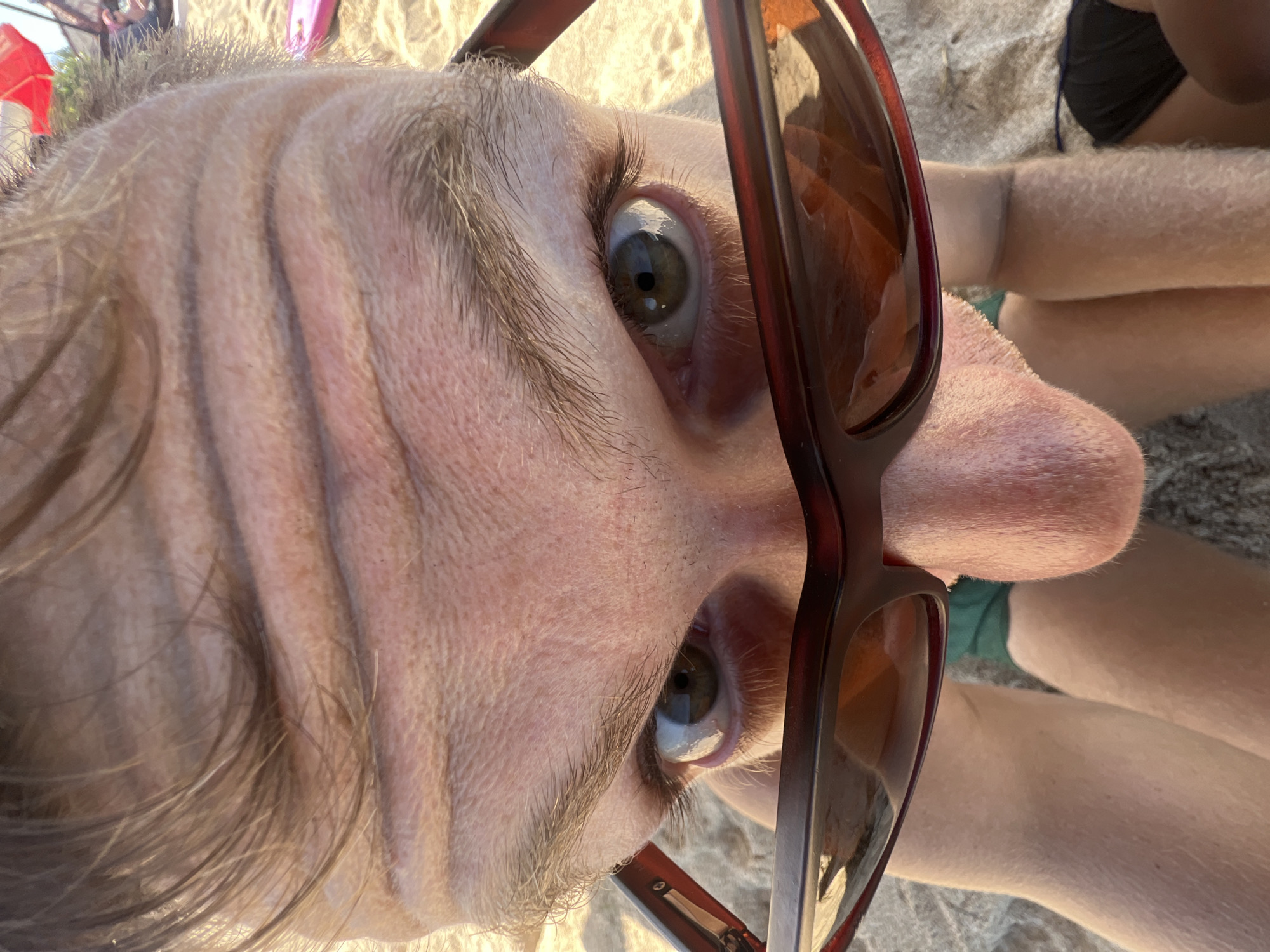 A close-up, low-angle shot shows a person's face from a beach perspective, with a dominant focus on the upper forehead and eyes, framed by sunglasses. The forehead displays prominent horizontal wrinkles. The individual wears sunglasses with dark, reflective lenses that obscure the eyes partially, revealing a portion of the irises. The nose is visible below the sunglasses, and the chin and upper lip area are also in frame. The subject's hair is short and light brown. The background suggests a beach environment, with sand visible in the lower portion of the frame, and elements like a red umbrella and possibly beach chairs or towels in the distance. The scene is lit by natural sunlight, casting a warm glow. The location is Kuta Selatan, Indonesia.