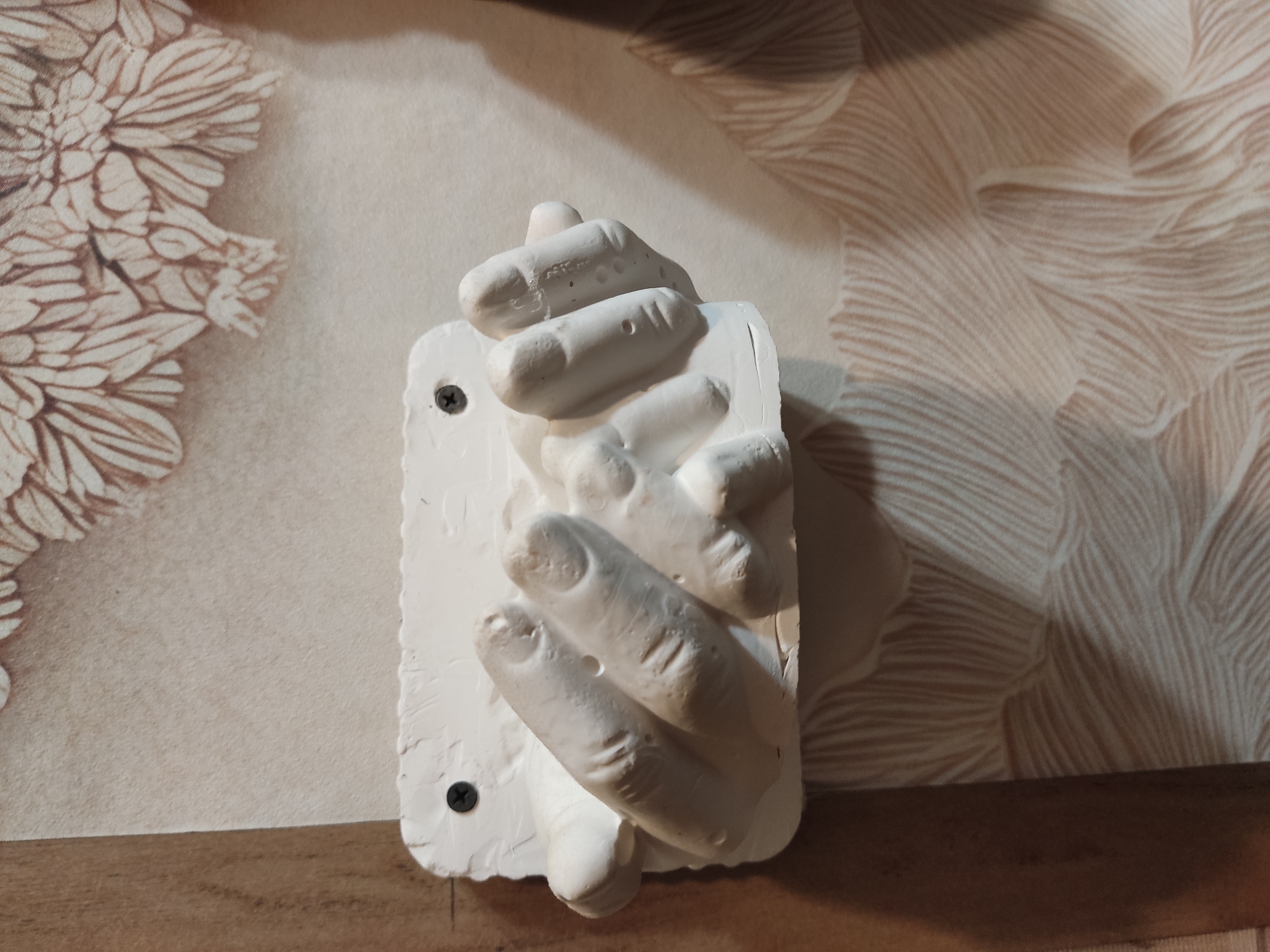 The image shows a close-up view of a white plaster cast of hands, mounted on a wall with a textured wallpaper featuring a floral pattern and some abstract lines. Two screws are visible securing the plaster cast to the wall. The cast appears to depict two hands clasped together. The lighting suggests an indoor environment, possibly a home or a decorative space. There is no information available in the image to determine its location in the city of Noginsk, Russia.