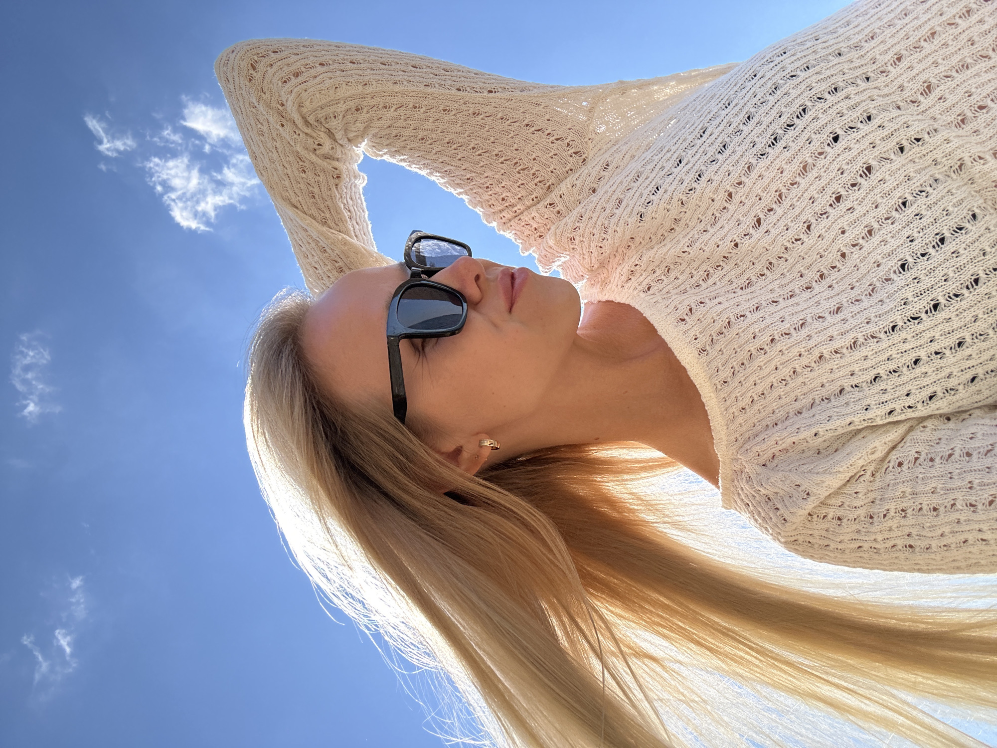 A blonde woman wearing sunglasses and a cream-colored, crocheted sweater stands outdoors against a bright blue sky with a few wispy clouds. Her left arm is raised, with her hand resting on the back of her head. Her long blonde hair is blowing to the left, illuminated by the sun. The image appears to be taken from a low angle, looking up at the woman. The location is stated to be Gornyi Erik, Russia.