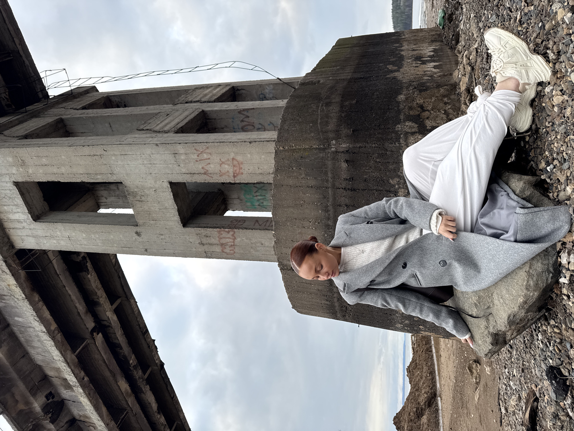 A full-body shot features a young woman seated outdoors on a large, dark grey rock. She is dressed in a light-colored sweater, white pants, a long grey coat, and chunky white sneakers. Her hair is pulled back, and her head is tilted downwards, suggesting a moment of contemplation or rest.
The setting is an industrial or abandoned area in Chaikovsky, Russia. Immediately behind the woman stands a massive, weathered concrete structure, possibly part of a former bridge or industrial complex. This structure has multiple rectangular openings and shows significant signs of age, including dark stains, crumbling sections, and visible rebar. Graffiti in red and green spray paint, with discernible words like "MIK" and "MIX," is present on the concrete beams.
The ground surrounding the woman and the structure is covered with a mix of small and large grey and brown pebbles and rocks. In the background, a calm, grey body of water, likely a river or reservoir near Chaikovsky, meets a distant, low-lying landmass. The sky is overcast, providing soft, diffused lighting, indicating a cloudy day. The overall atmosphere is stark and quiet, blending elements of urban decay with the natural, rocky shoreline. The image is composed with the large concrete structure dominating the left side, providing a strong vertical and diagonal element against the horizontal expanse of the ground and water.