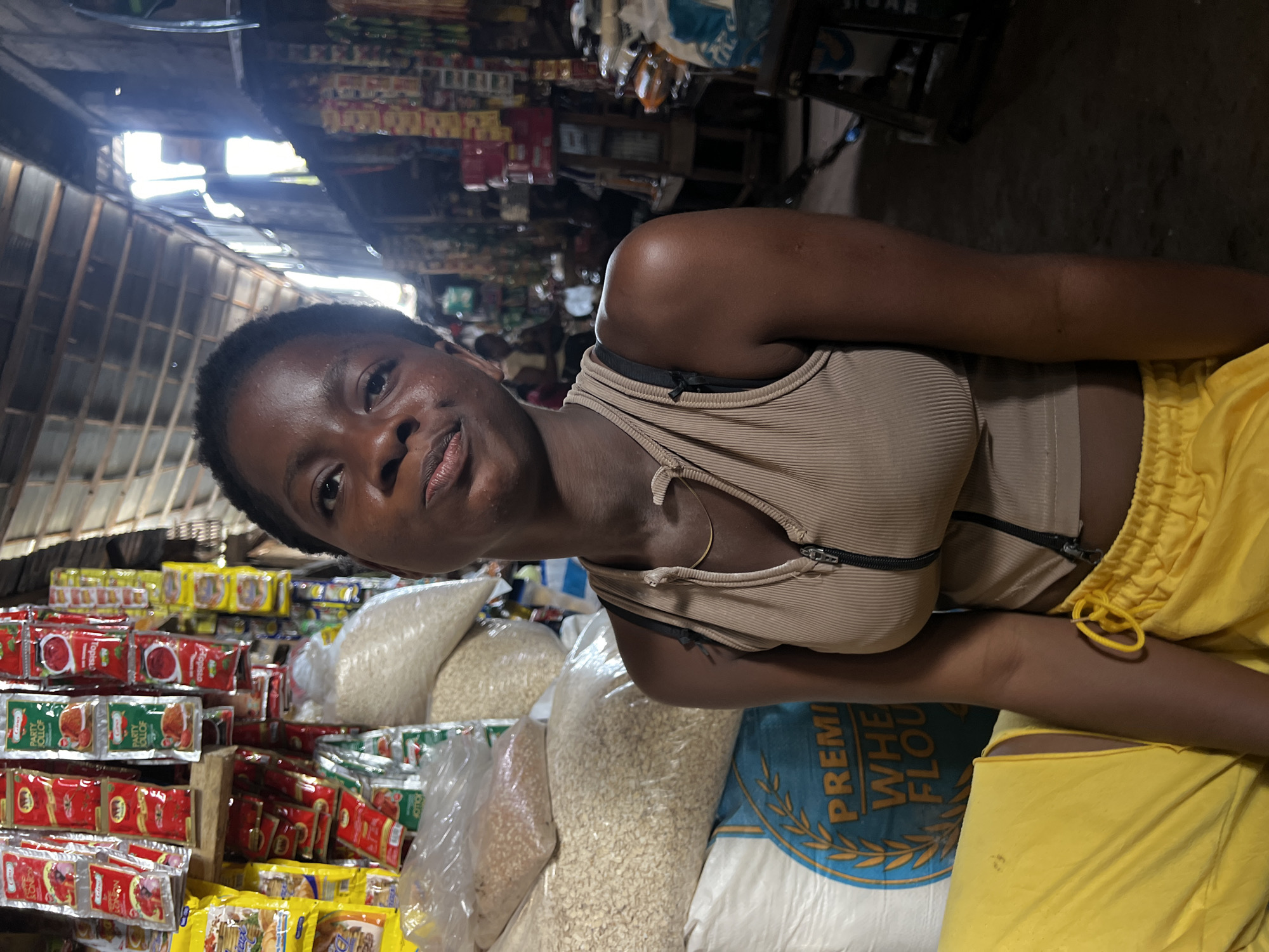 In Nkpor, Nigeria, a young Black woman is photographed from the chest up, seated in what appears to be a market stall. She has short, dark hair and is looking directly at the camera with a neutral expression. She is wearing a beige, ribbed tank top with a zipper down the front and yellow drawstring shorts. Behind her, the market is stocked with various goods. To her left, large sacks of what appears to be grain or flour are stacked high, with one sack clearly labeled "PREMIUM WHEAT FLOUR" in blue and yellow lettering. To her right, shelves are filled with packaged food items, including brightly colored sachets of spices and sauces. The market setting is rustic, with a corrugated metal roof visible overhead and natural light filtering in from the open sides. In the background, other market-goers are visible, suggesting a bustling commercial environment.