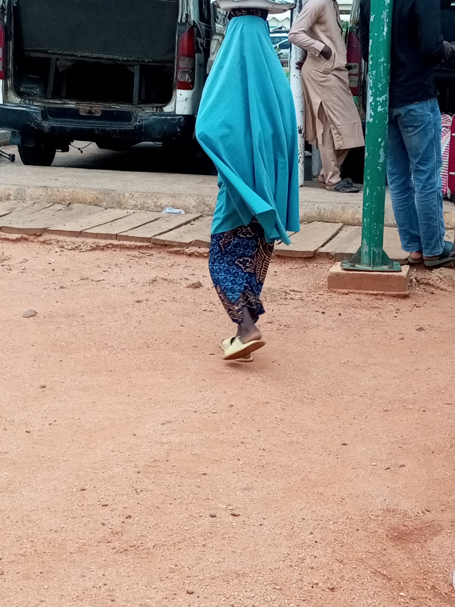The image depicts a scene on a dirt road in the city of Gombe, Nigeria. In the foreground, a person is walking away from the camera, wearing a vibrant turquoise hijab that drapes over their body and a patterned blue and white skirt. They are also wearing light-colored sandals. To the left of the person, the rear of a white van is visible, appearing to be parked. The van's doors are open, revealing a dark interior. To the right, a green metal pole stands on a concrete base, and beyond that, two other individuals are standing. One person, on the far right, is wearing blue jeans and dark sandals, with their back to the camera. Next to them, a person in a light beige robe with their hands in their pockets, also facing away from the camera, is leaning against a surface. The ground is a dusty, reddish-brown earth mixed with small pebbles. Wooden planks are laid out on the side of the road.